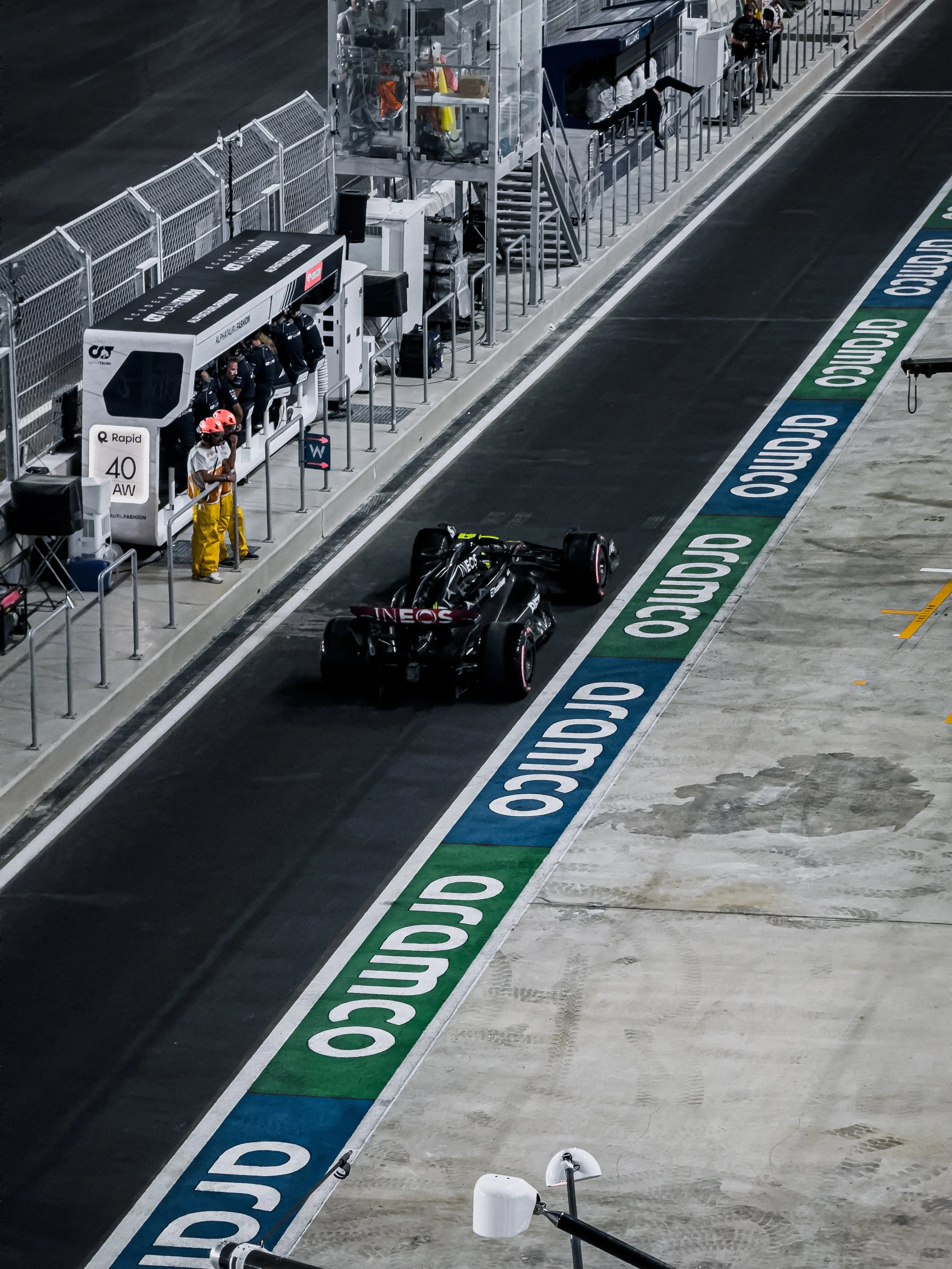 A racing car is in the pit lane of a race track, near the team's garage and pit crew. The pit lane is marked with blue and green lines with the word 'OMICO', and the scene is illuminated at night.