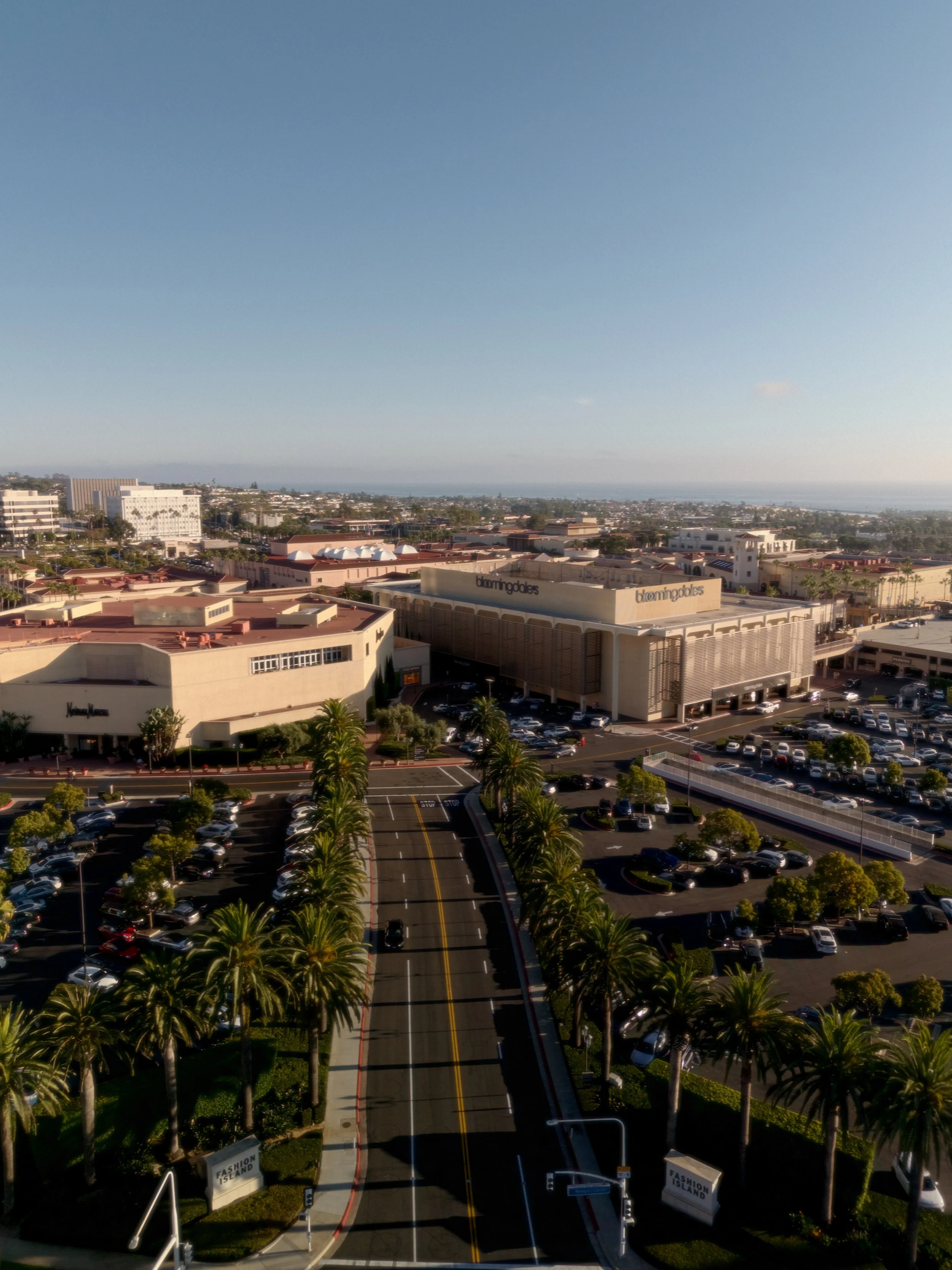 Aerial view of a shopping mall parking lot with palm trees, cars, and buildings labeled Bloomingdale's and Nordstrom in a coastal city.