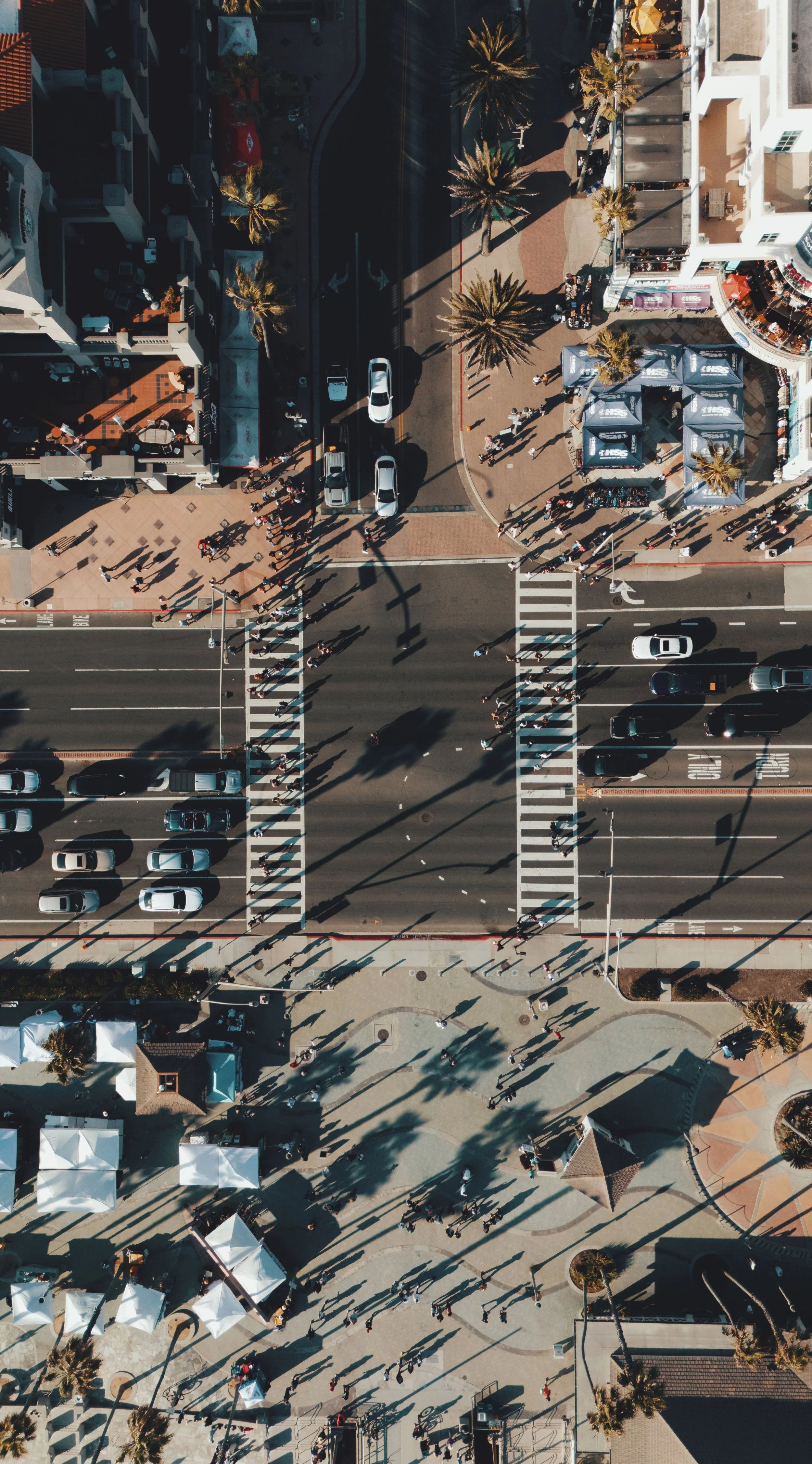 Aerial view of a busy street intersection with cars, pedestrians, palm trees, and outdoor tents and booths.