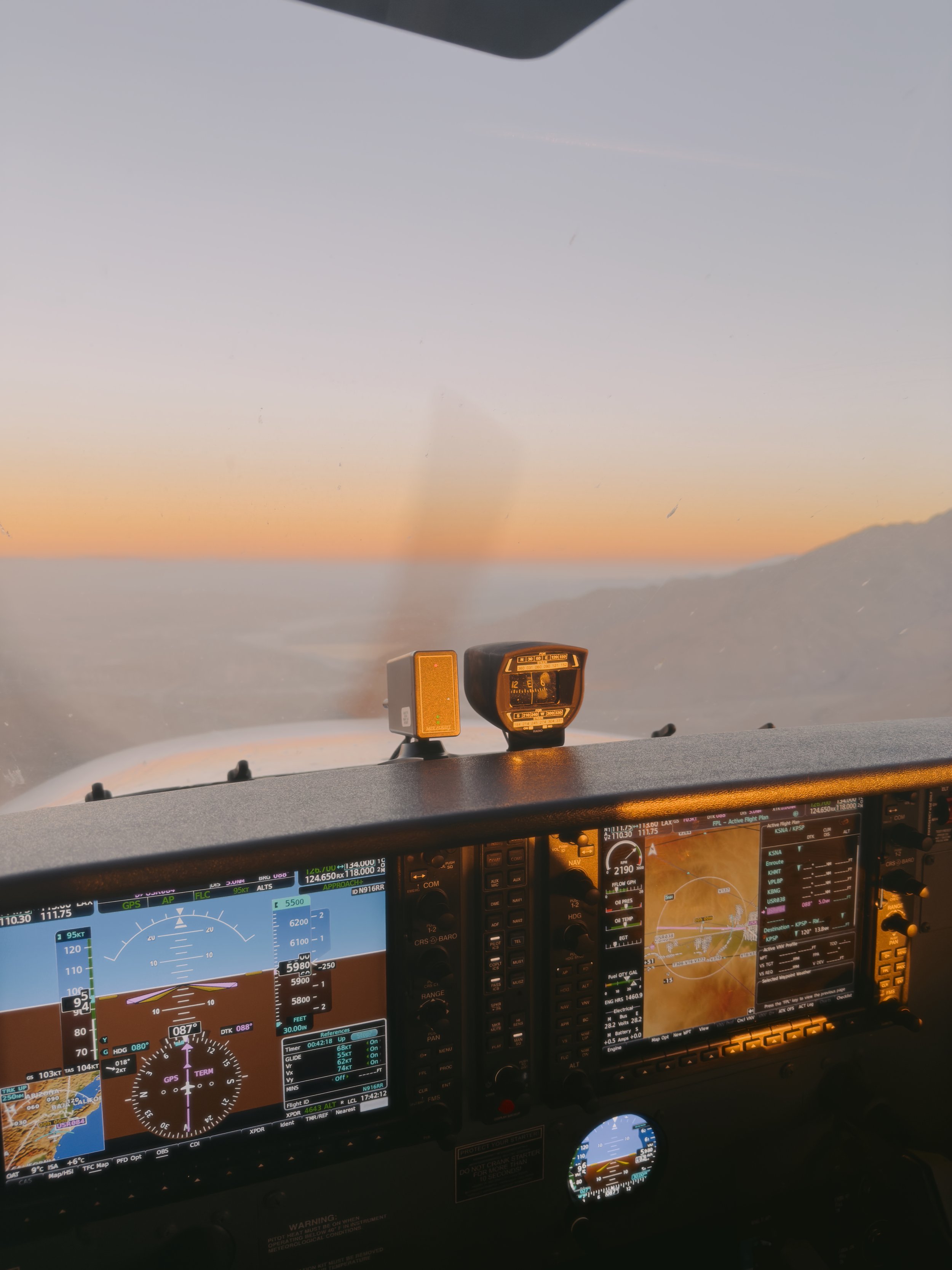 Inside an airplane cockpit during sunset, showing multiple flight instruments and displays with a view of the sky and landscape outside the window.