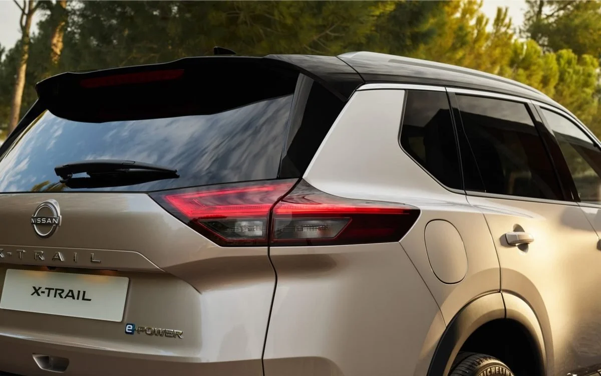 Auto Nissan X-Trail di colore beige con luci posteriori a LED, in un'ambientazione naturale con alberi e cielo riflessi sul vetro posteriore.