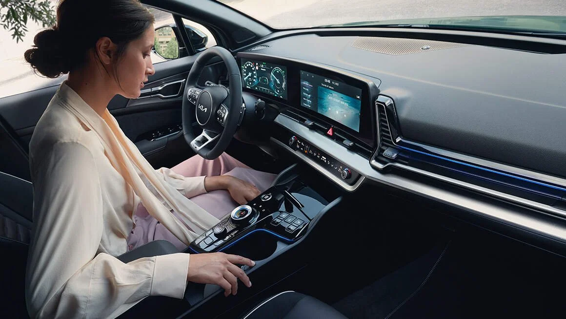 Interno moderno di un'auto con una donna seduta alla guida che interagisce con il cruscotto digitale e i display touchscreen.
