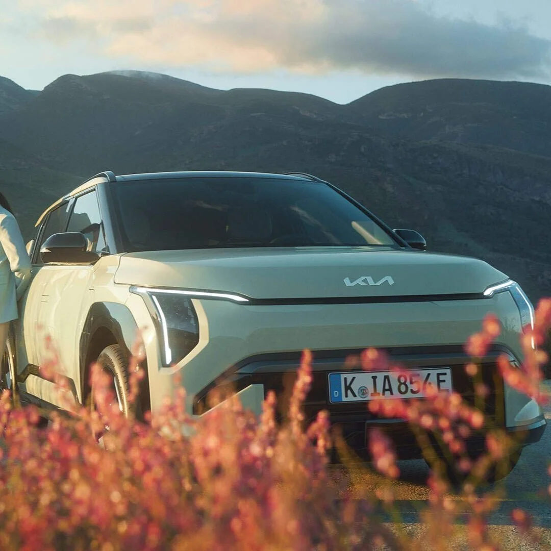 Auto elettrica Kia su strada con montagne sullo sfondo e fiori in primo piano.