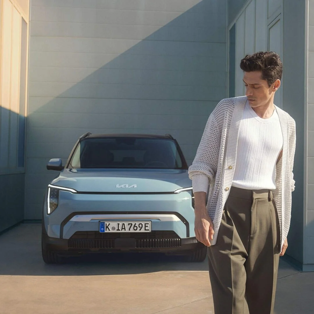 Uomo vestito eleganti in piedi davanti a un'auto elettrica di colore azzurro, si trova su un vialetto accanto a un edificio moderno