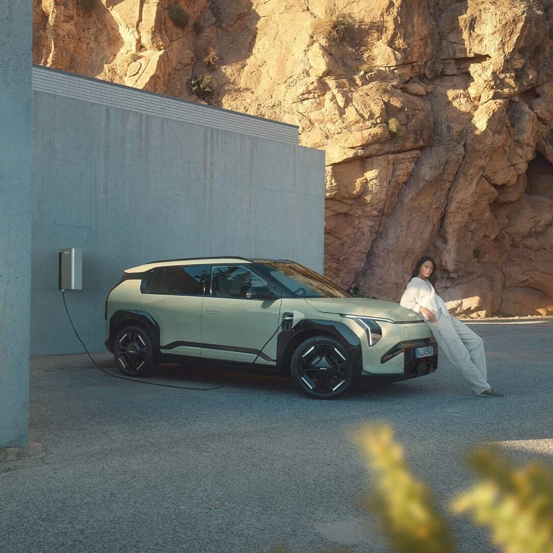 Una donna seduta accanto a un'auto elettrica collegata a una stazione di ricarica, con uno sfondo roccioso e un muro grigio.