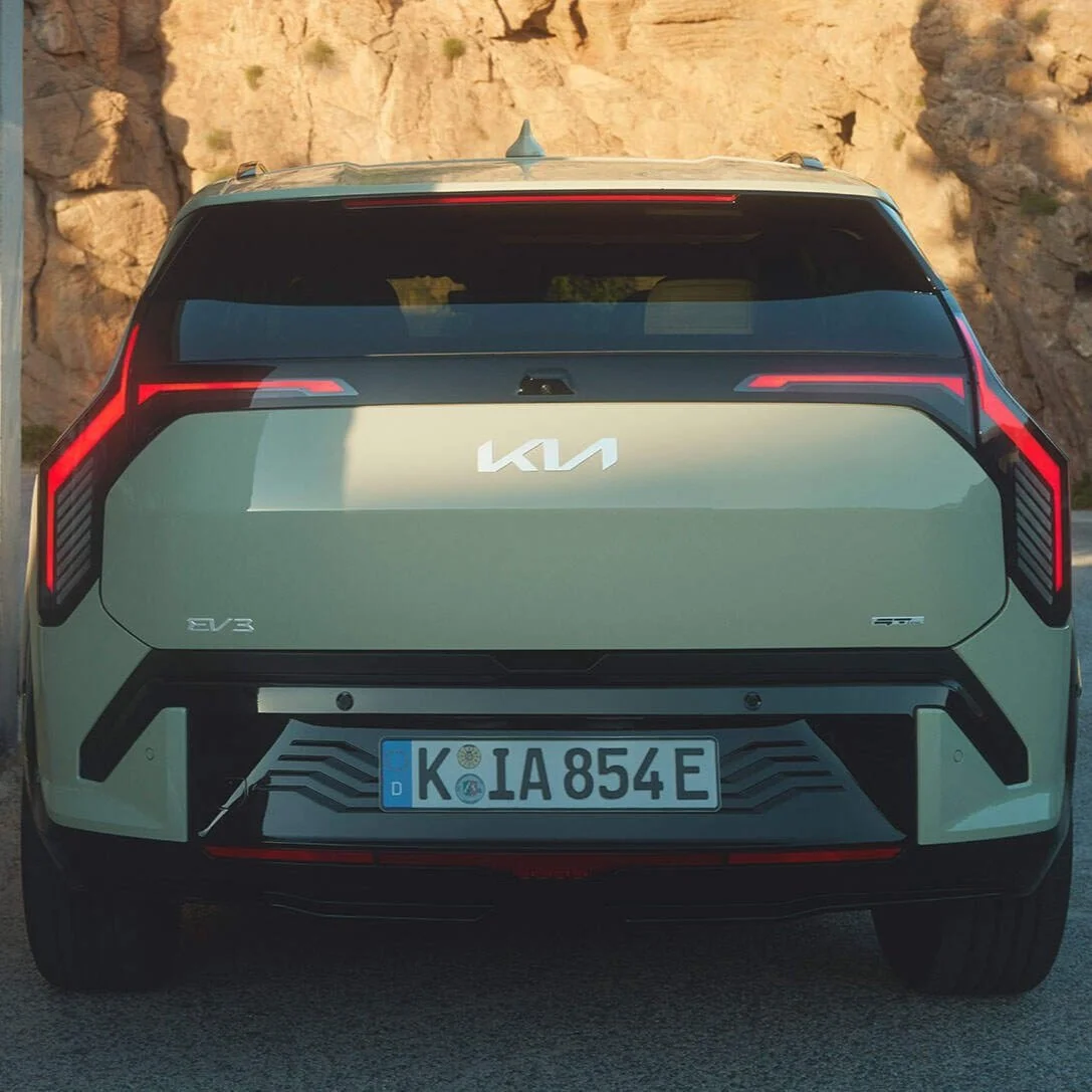 La parte posteriore di un'auto Kia EV3 di colore grigio, con luci posteriori rosse e targa tedesca, in un paesaggio di rocce e natura.