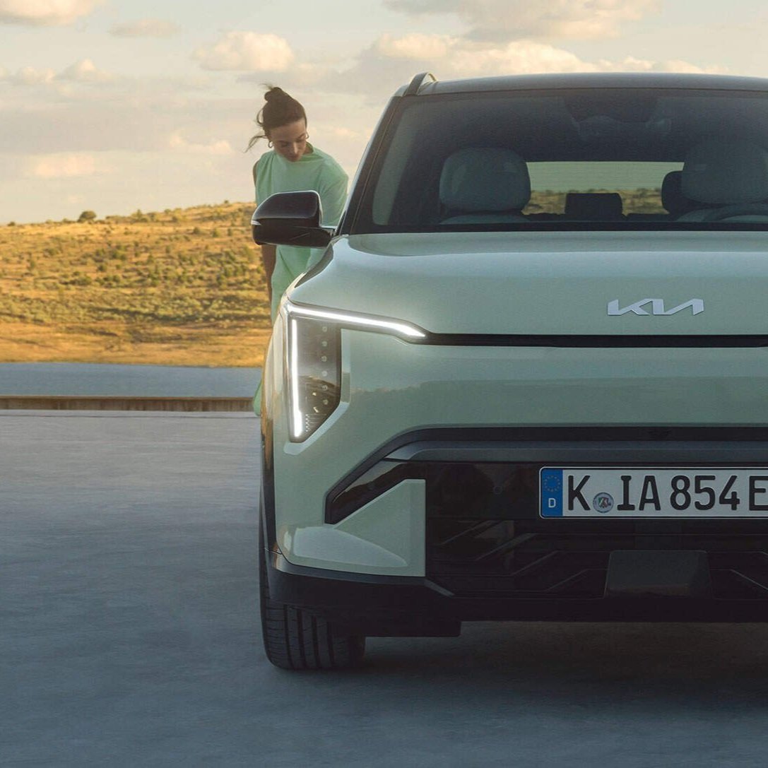 Auto elettrica bianca KIA sulla strada con una donna che si sta avvicinando, sfondo di colline verdi e cielo nuvoloso