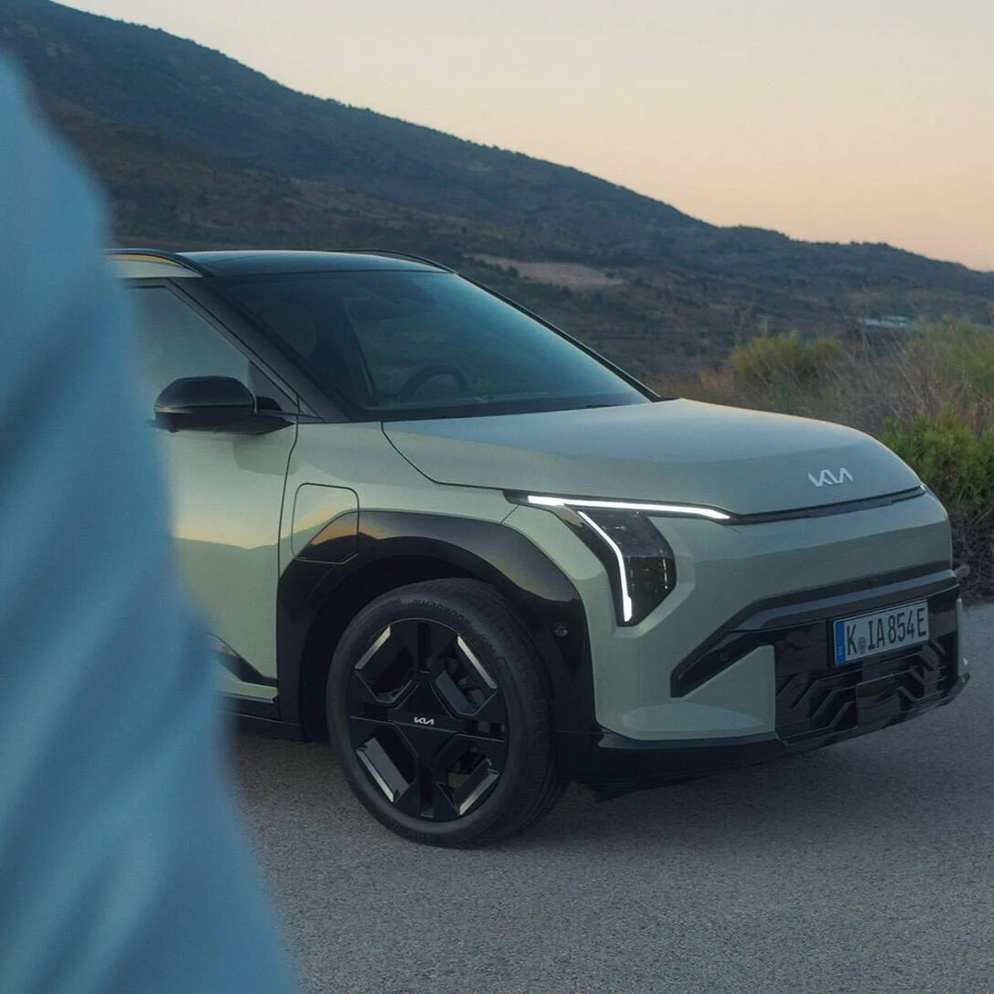 Un'auto elettrica di colore verde chiaro si trova su una strada con uno sfondo di colline e cielo al tramonto.