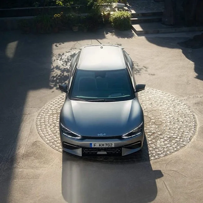 Auto grigia vista dall'alto in un cortile, con ombra proiettata davanti e piante sullo sfondo.