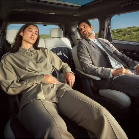 Due persone, una donna e un uomo, seduti in un'auto, sembra che siano in viaggio, con paesaggi verdi visibili attraverso i finestrini.