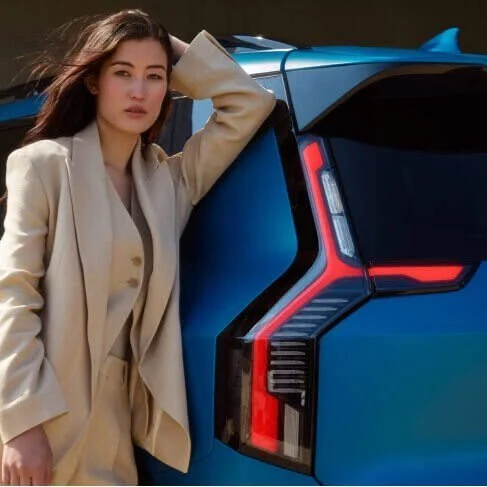 Una donna con un abito beige che si appoggia a una vettura blu con luci a LED rosse sul retro.