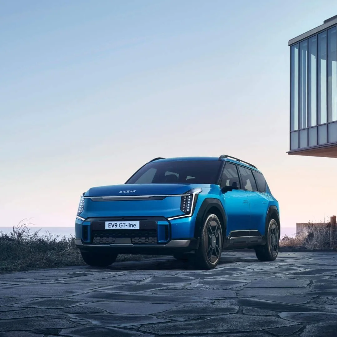 Auto blu Kia EV9 GT-line parcheggiata su una strada di pietra con edifici moderni sullo sfondo al tramonto.