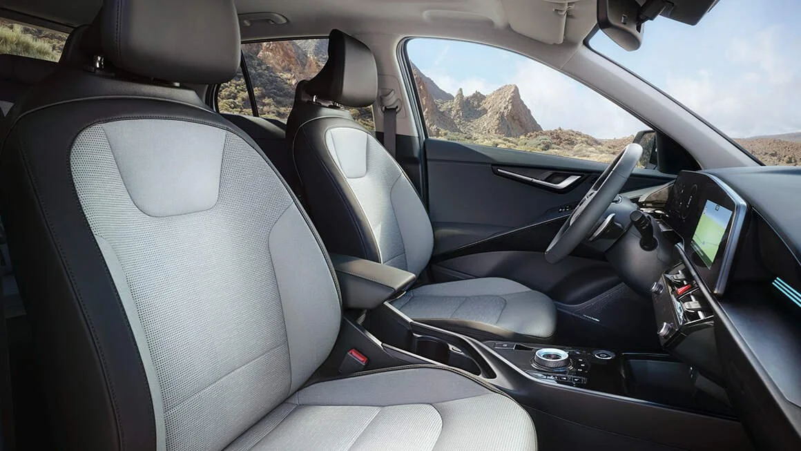 Interno di un'auto moderna con sedili grigi e schermo digitale, con un paesaggio montano visibile attraverso il parabrezza.