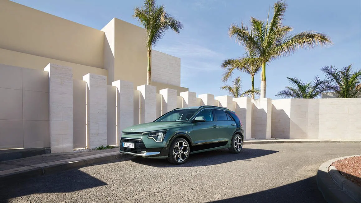 Auto verde scuro parcheggiato su strada davanti a un muro bianco con palme sullo sfondo e un cielo chiaro.