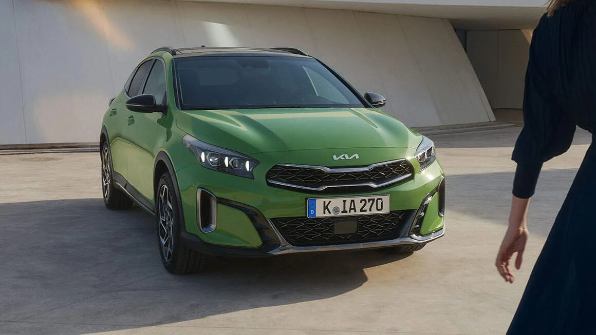Auto Kia di colore verde con targa europea, parcheggio all'aperto, persona con vestito blu che si avvicina.