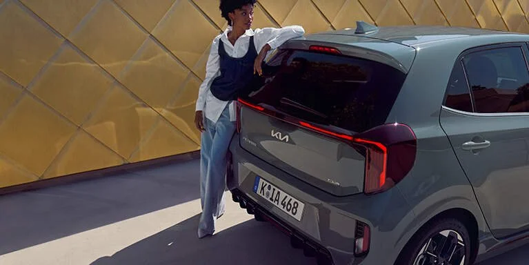 Donna che si appoggia a una vettura Kia grigia in un'area con muro giallo decorato