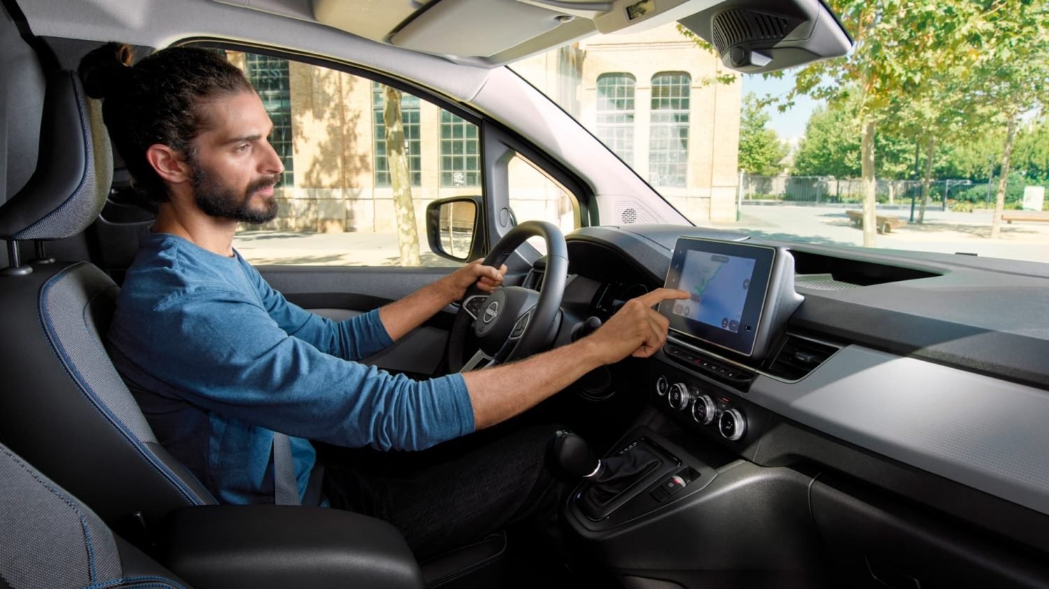 Uomo con capelli raccolti al galera alla guida di un'auto, toccando uno schermo di navigazione sul cruscotto in una giornata soleggiata con alberi e edifici sullo sfondo.