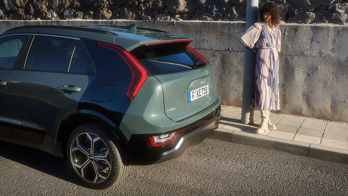 Una donna con un vestito lungo a righe e stivali bianchi si appoggia a un palo della luce sulla strada, di fianco a un'auto di colore verde scuro.