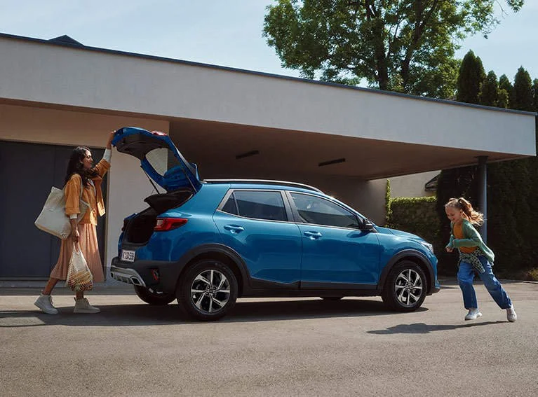 Due donne aprono il portellone di un'auto blu con una valigia e una borsa, mentre una giovane ragazza corre verso di loro, in un'area di parcheggio vicino a un edificio moderno con un albero in background.