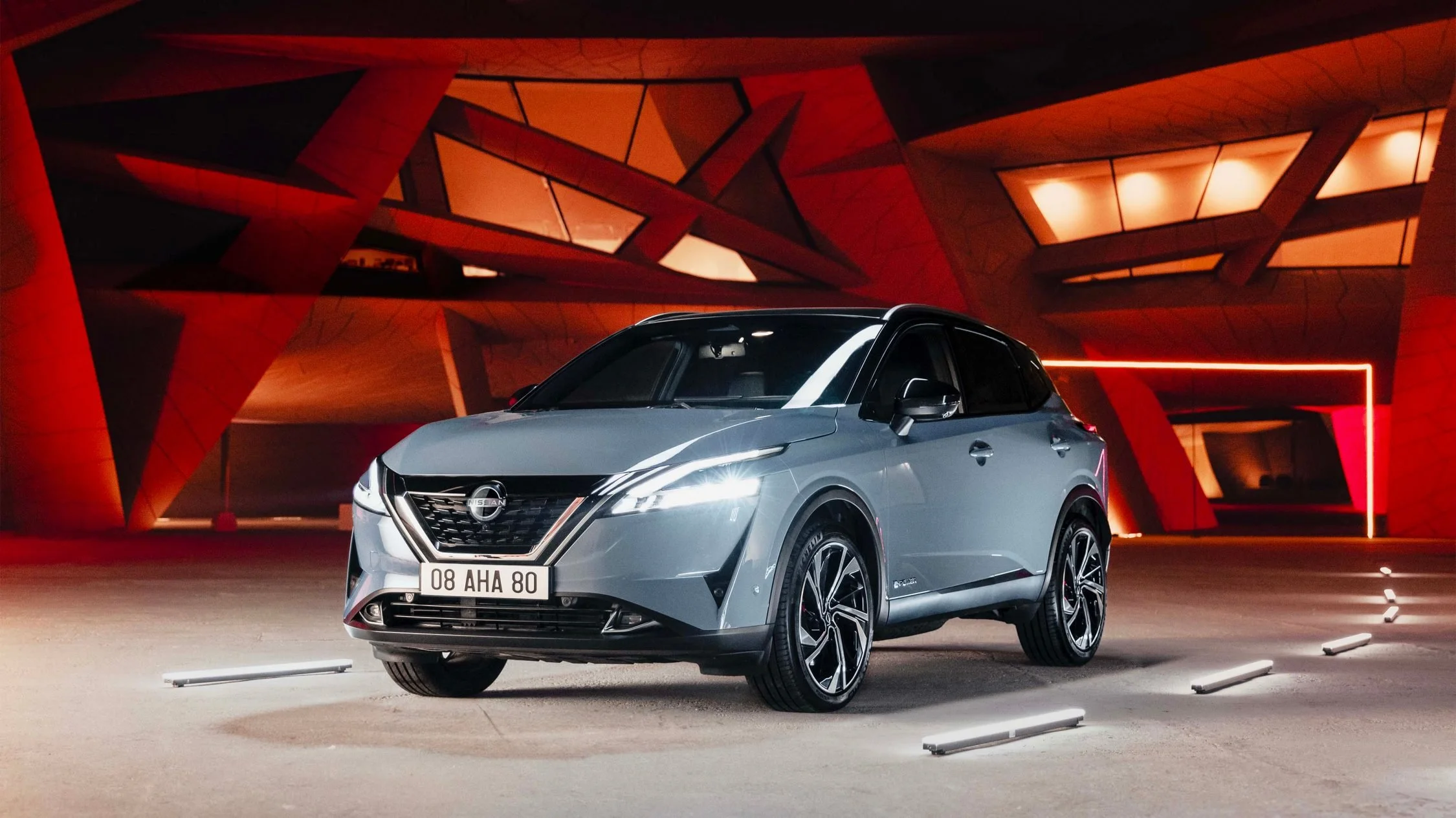 Auto Nissan di colore grigio chiaro con luci a led anteriori, parcheggiata in un'area urbana notturna con architettura moderna in sfondo, illuminata da luci rosse e neon.