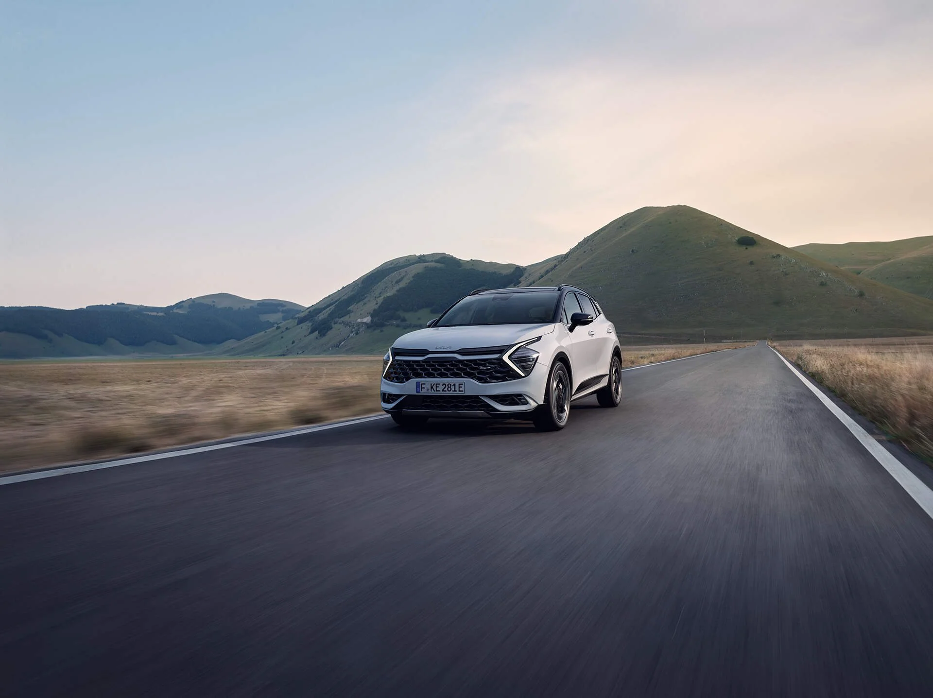 Una macchina moderna bianca in corsa su una strada di campagna con colline verdi sullo sfondo, al tramonto.