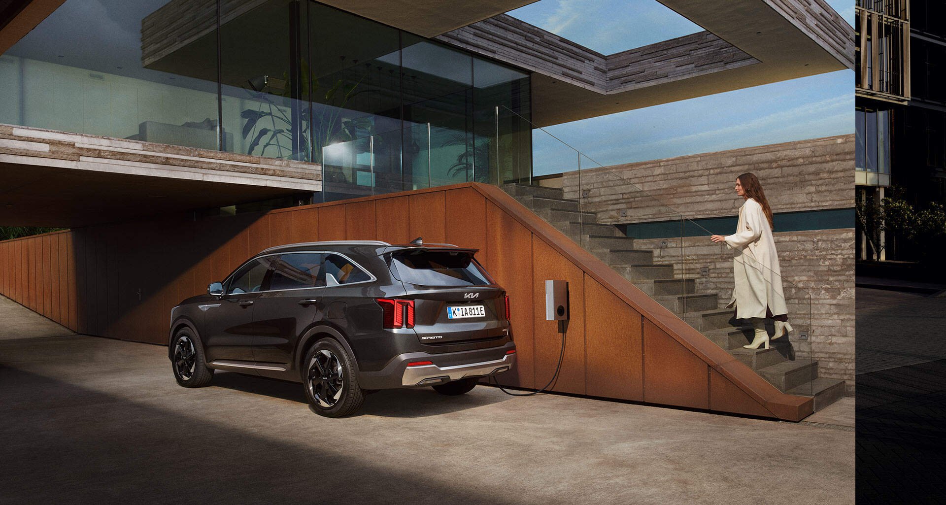 Automobile elettrica collegata a una stazione di ricarica accanto a una moderna residenza con scale esterne e pareti in pietra, con una donna che sale le scale in abiti chiari.