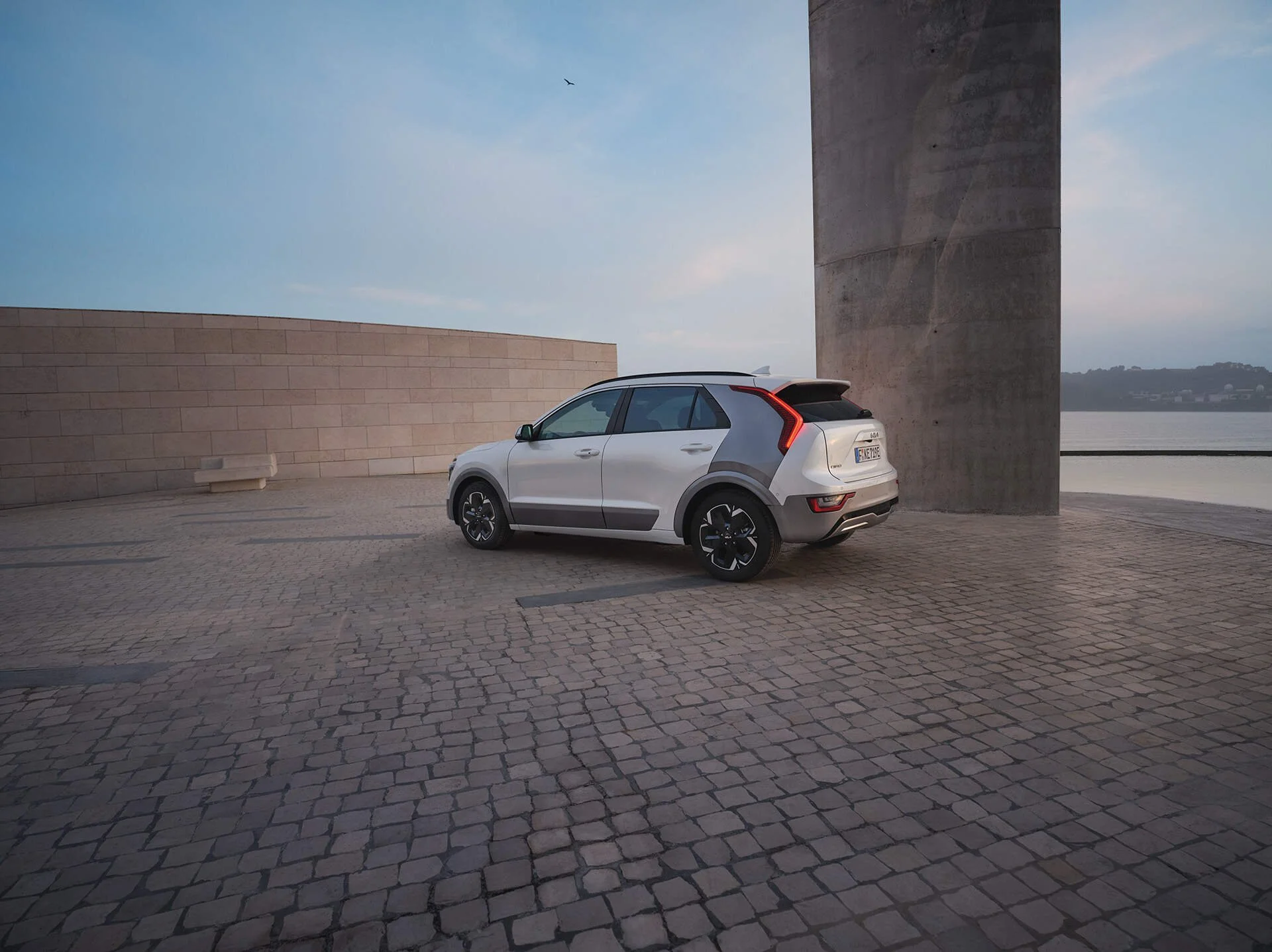 Auto elettrica grigia parcheggiata su una piazzale di pietra vicino a un grande muro di cemento e un fiume, con un cielo sereno e poca nuvolosità.
