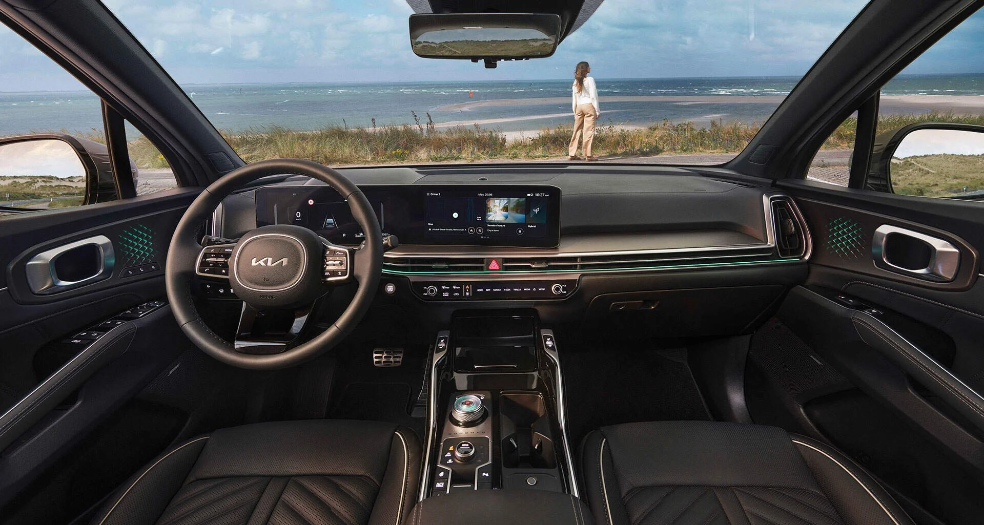Interno di un'auto moderna con display digitale, sedili neri e volante, vista sulla spiaggia e una donna che guarda l'oceano fuori dal finestrino.