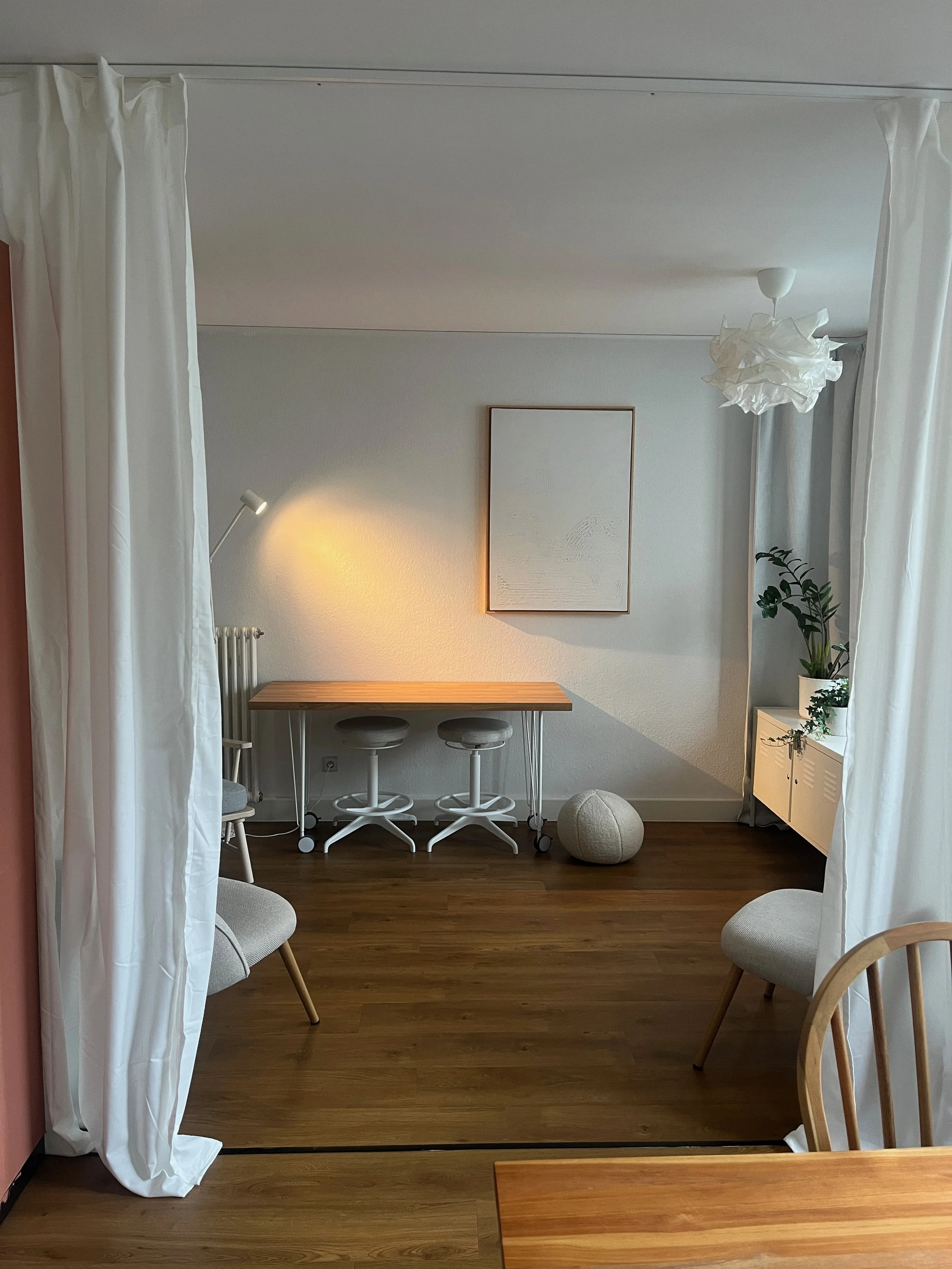 Ein heller Raum mit weißen Vorhängen, einem hohen weißen Lampenschirm, einem kleinen Wandbild, einem weißen Sideboard mit Pflanzen, einem Tisch und Stühlen, Holzboden und einem Stehlampenlicht.