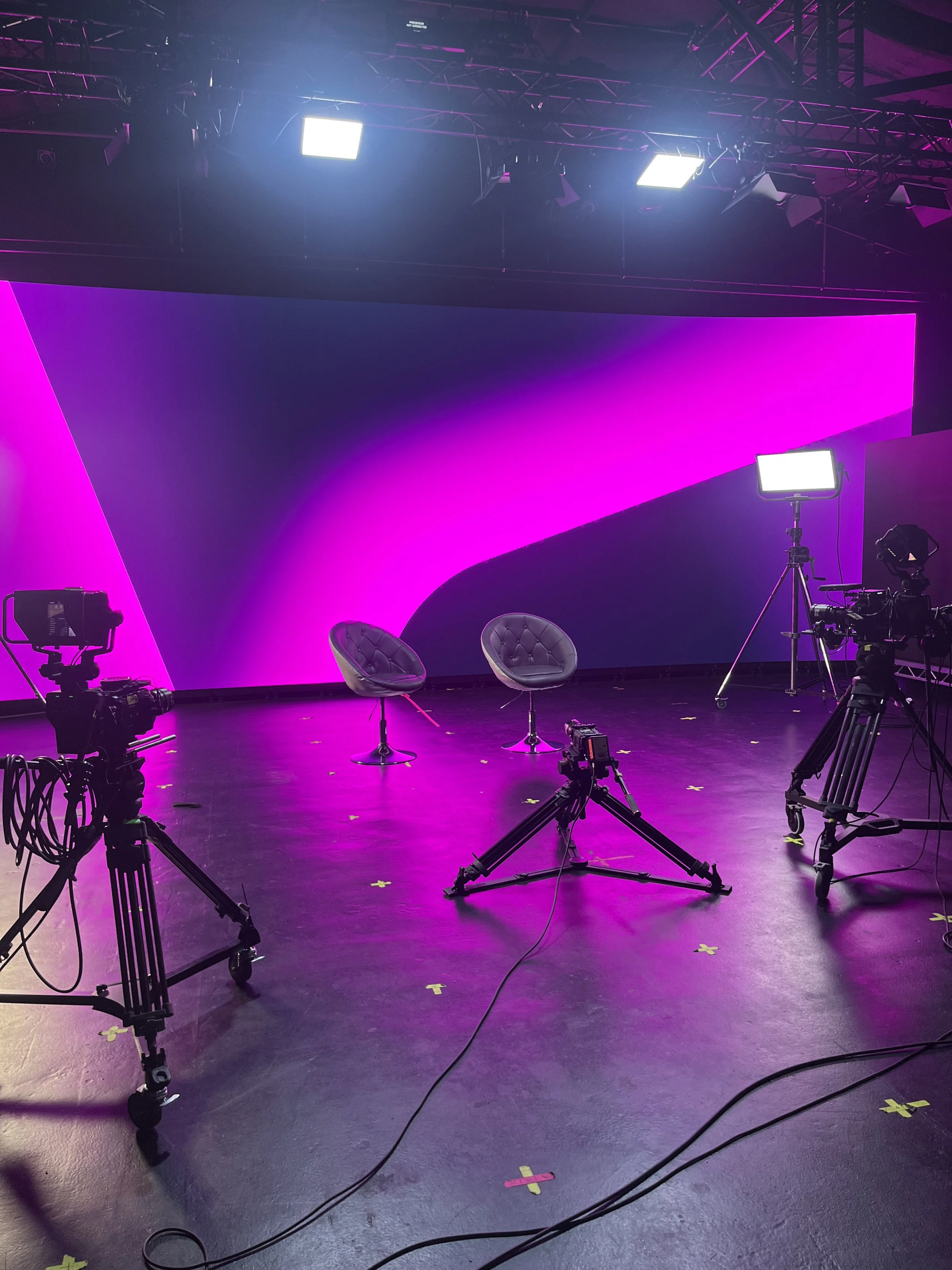 Empty studio setup prepared for a filmed conversation, featuring cameras, lighting, and chairs positioned in front of a curved LED wall with a coloured backdrop.