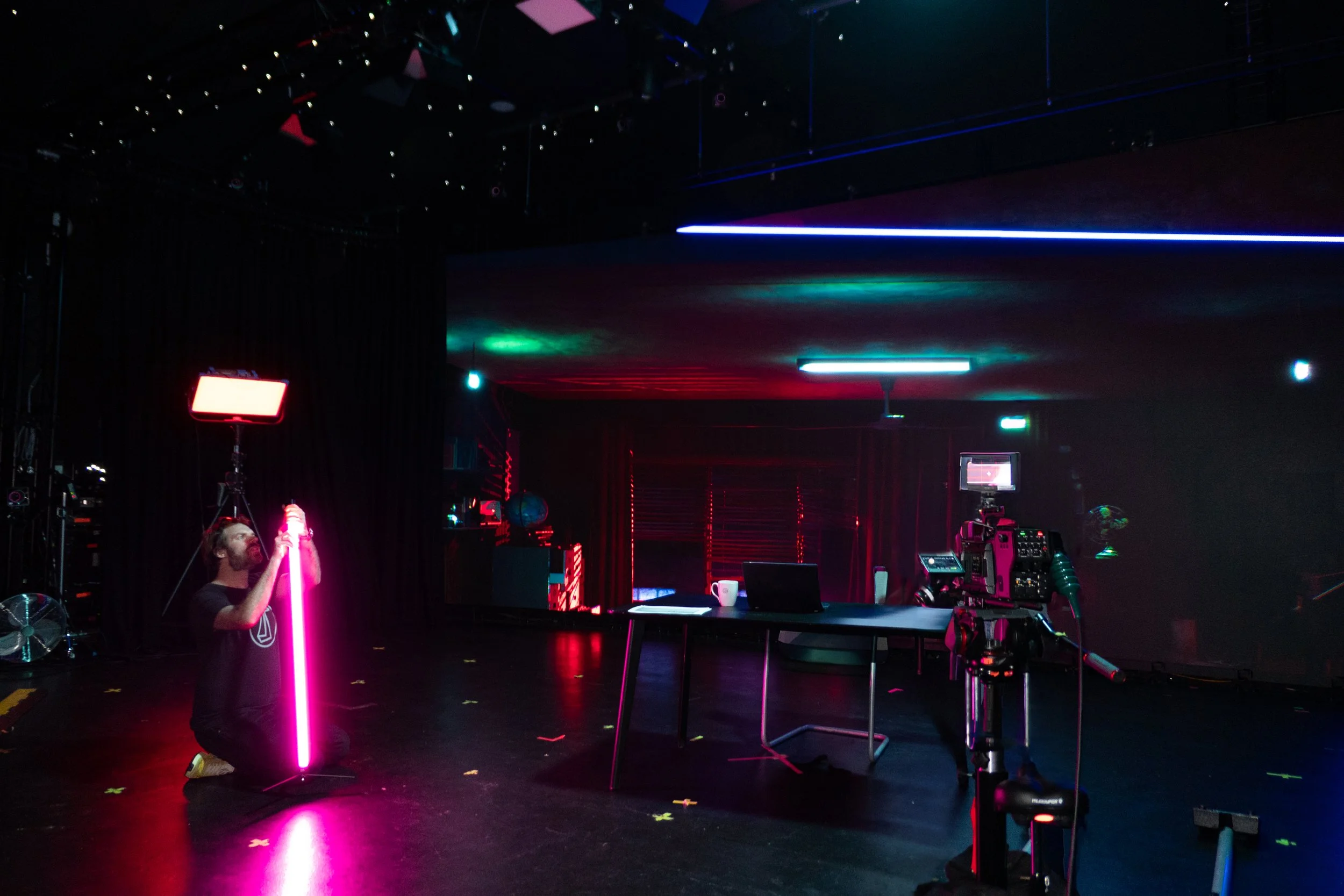 Behind-the-scenes view of a virtual production studio, showing camera rigs, lighting equipment, and crew adjusting a neon light element on a dark LED stage.