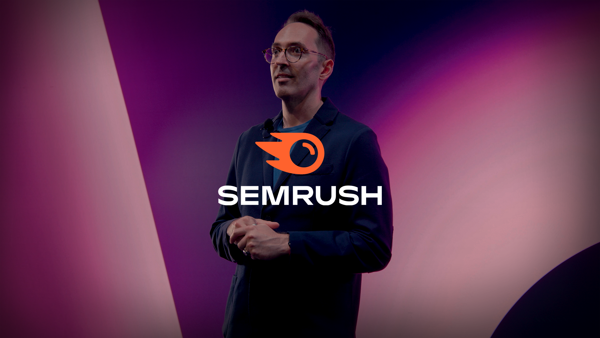 Speaker on set during a Semrush video production, standing against a branded gradient background with the Semrush logo centered, filmed in a studio environment.