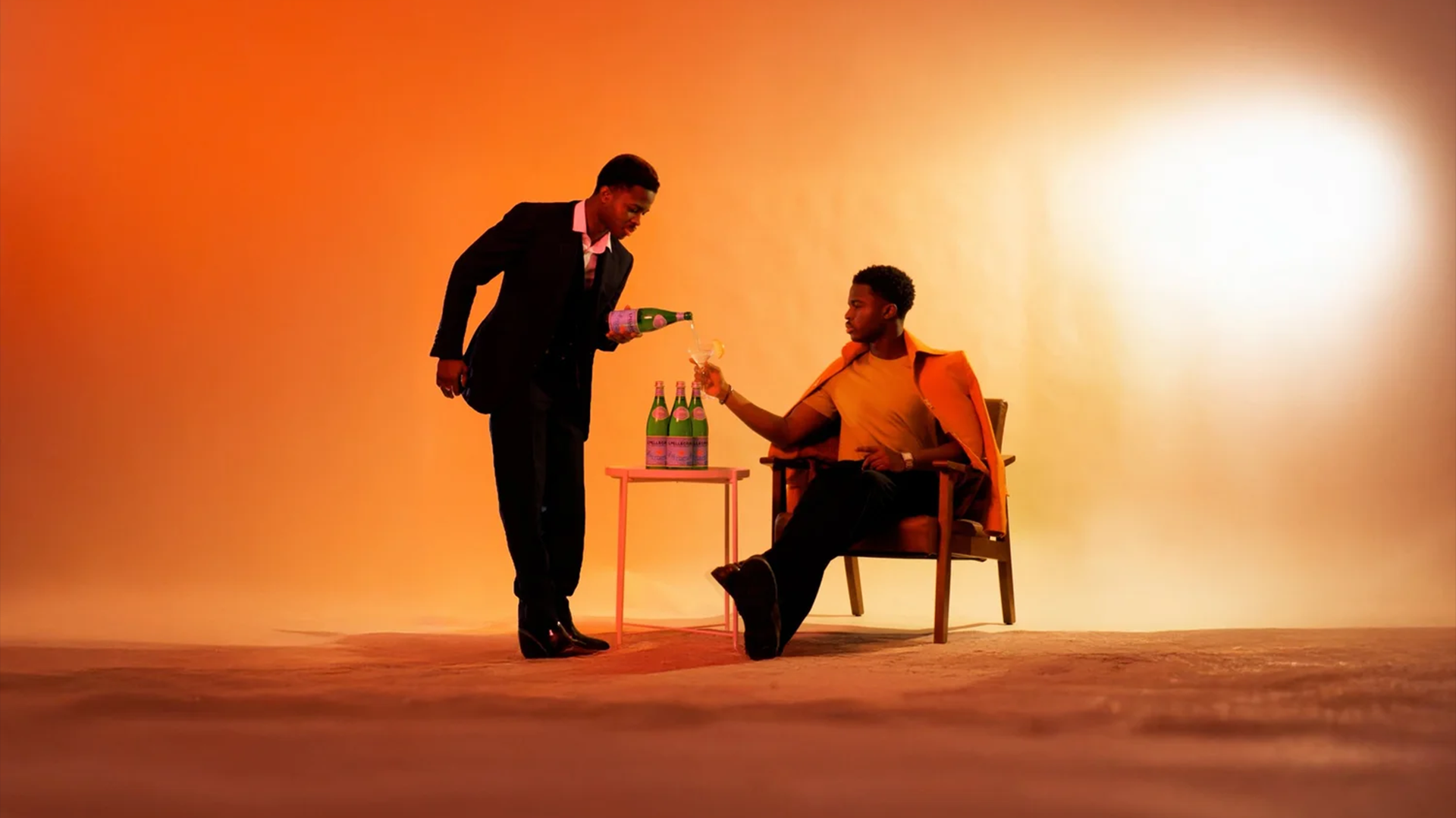 Still from a San Pellegrino spec commercial, showing two men on a stylised studio set as one pours sparkling water for the other, highlighting a VFX-led visual concept with warm, cinematic lighting.