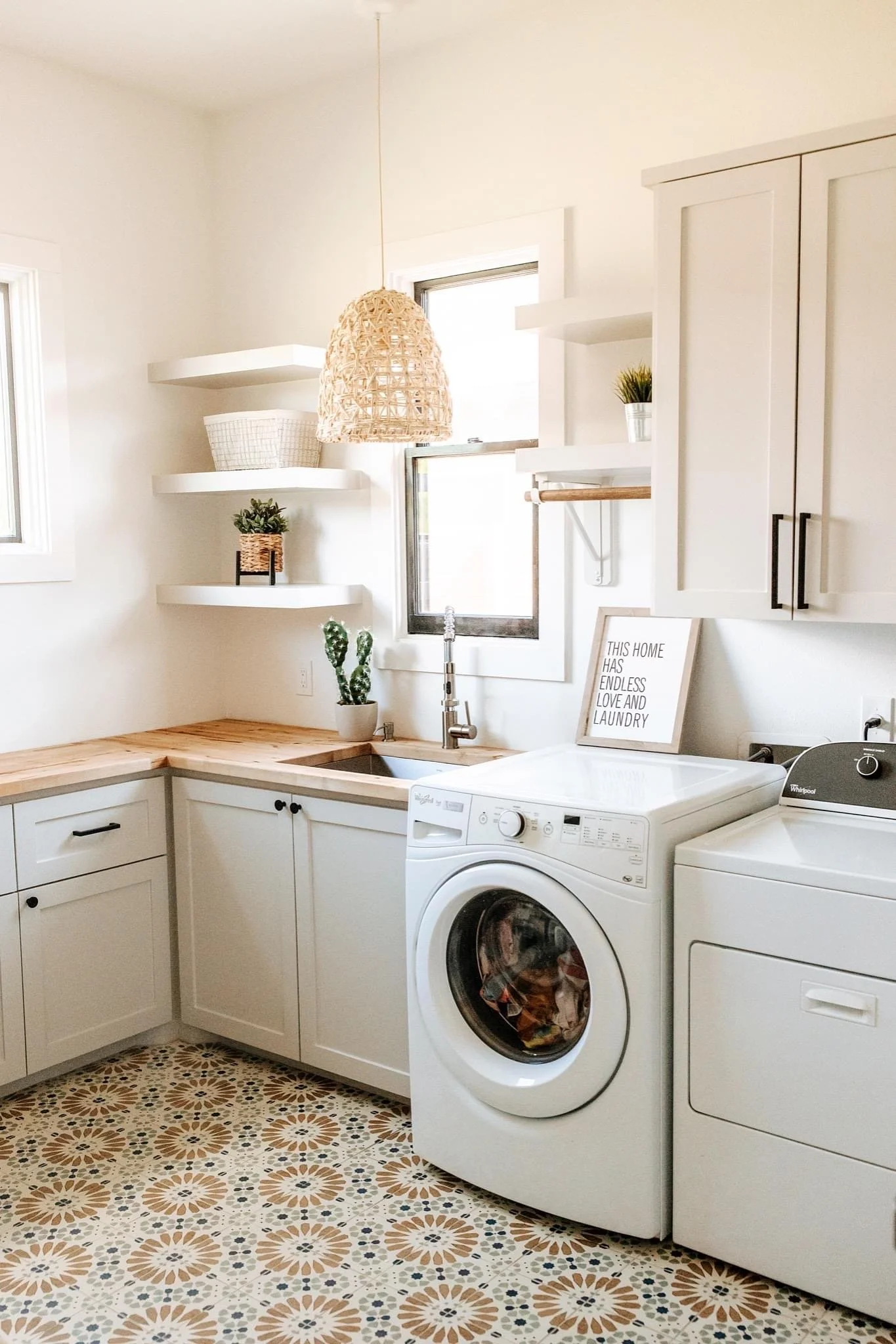 Laundry room with white cabinets, a washing machine, a dryer, patterned tile floor, wall shelves with decorative items, and a framed quote reading "This home has endless love and laundry."