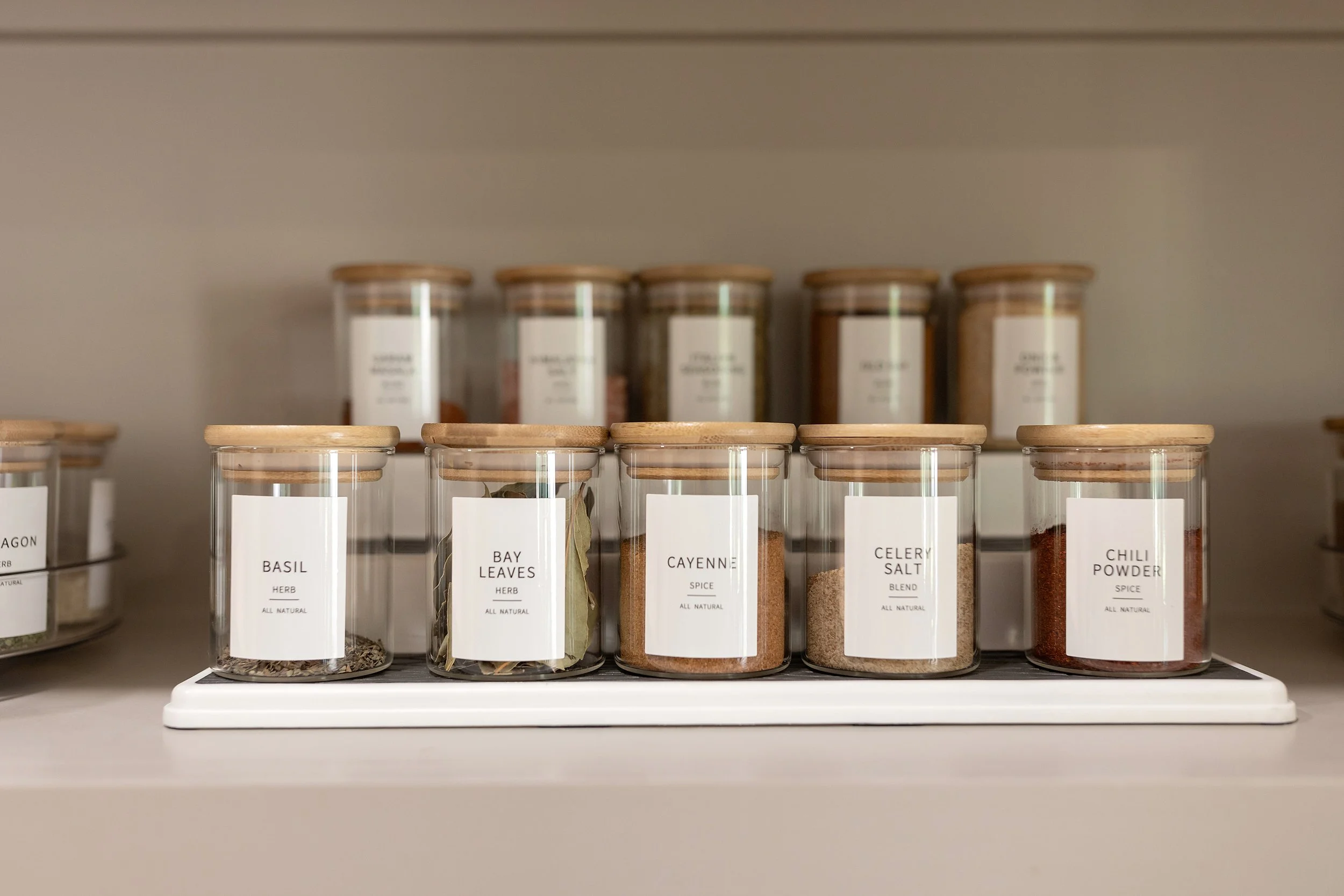 A shelf with glass jars containing herbs and spices, labeled Basil, Bay Leaves, Cayenne, Celery Salt, and Chili Powder.