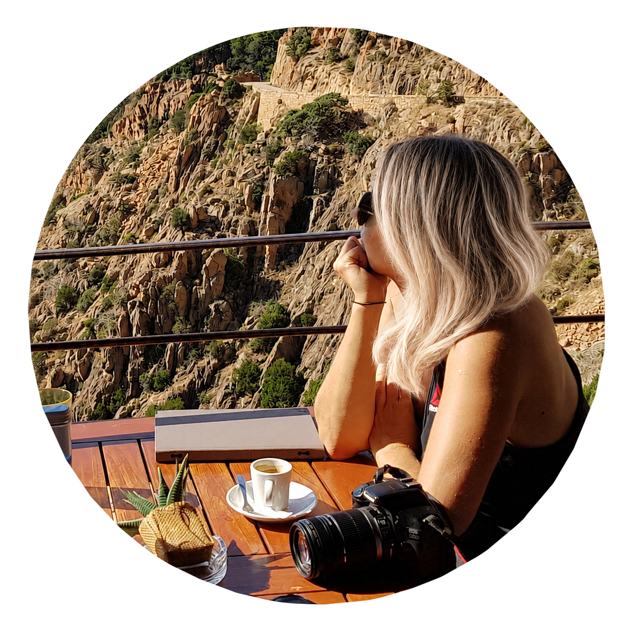 Laetitia Munduteguy with blonde hair and sunglasses sitting at a table, looking at a rocky mountain landscape with trees, coffee, a camera, and a small plant on the table in Corsica.