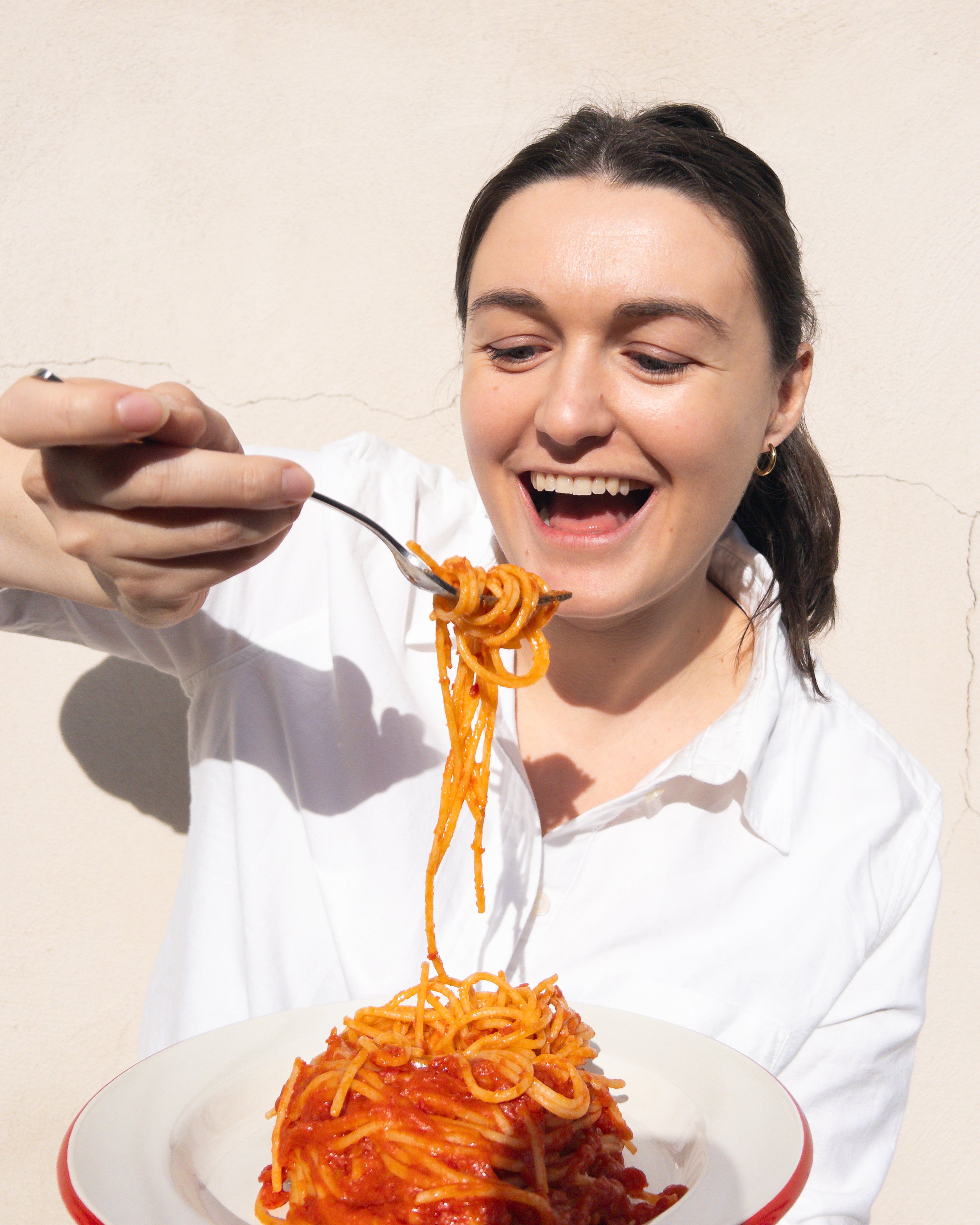 A smiling woman in a white shirt is holding a fork with spaghetti and tomato sauce, about to eat it. The spaghetti is on a white plate with a red rim.