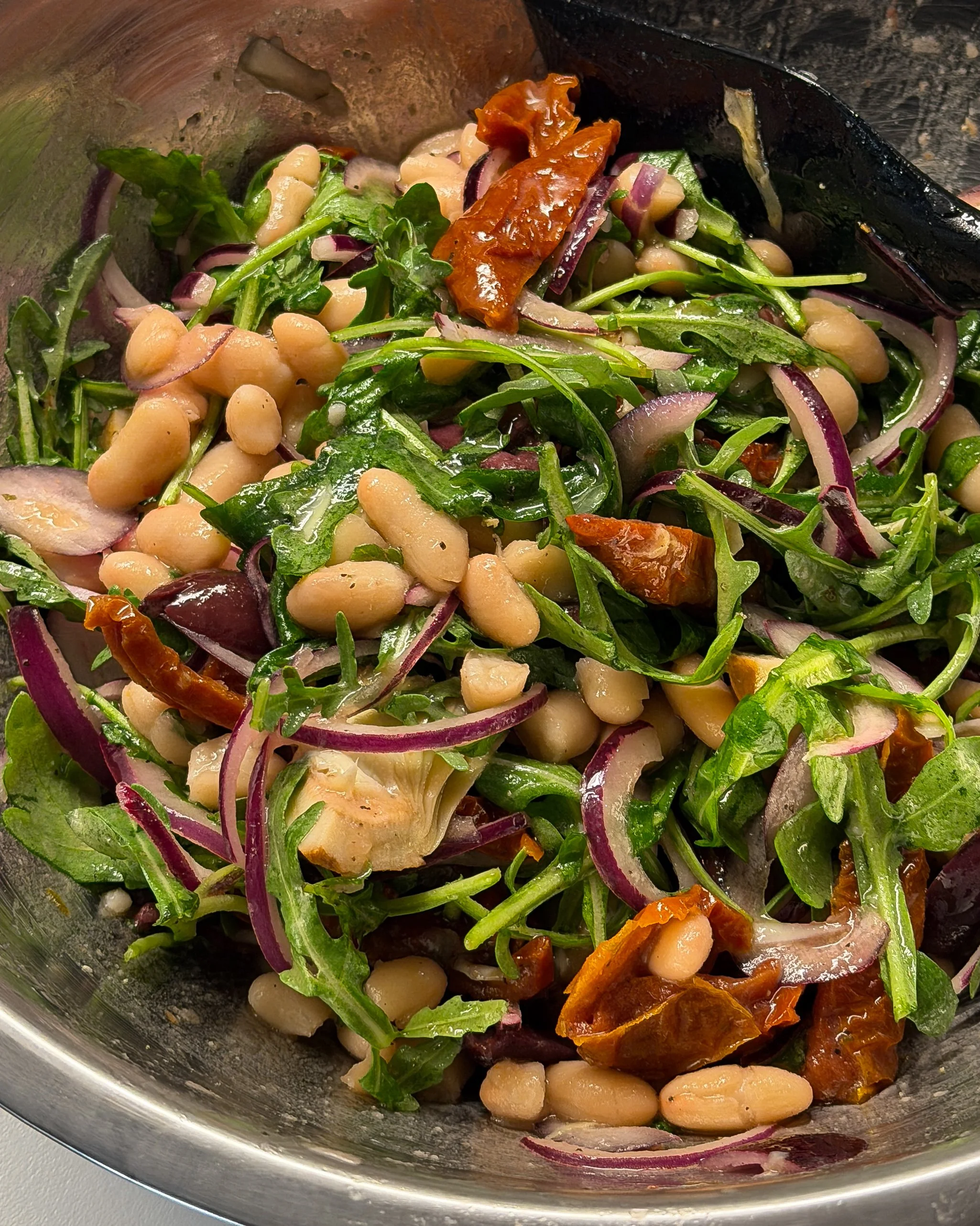 Marinated dense bean salad with cannellini beans, arugula, red onion slices, sun-dried tomatoes, and artichoke hearts in a metal bowl.