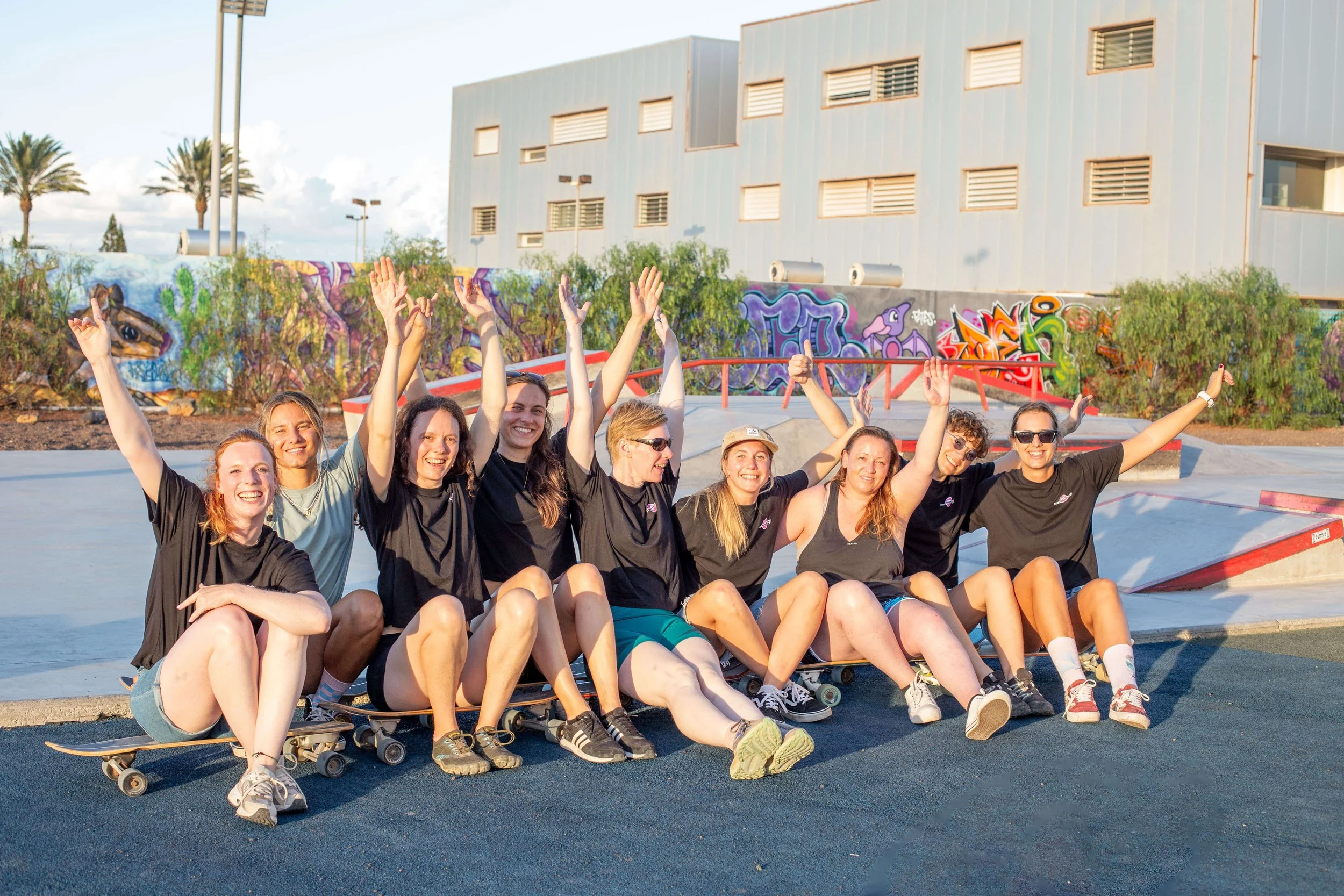 Group of young women sitting on skateboards at a skate park, smiling with arms raised in celebration, graffiti on the back wall, modern building in the background.