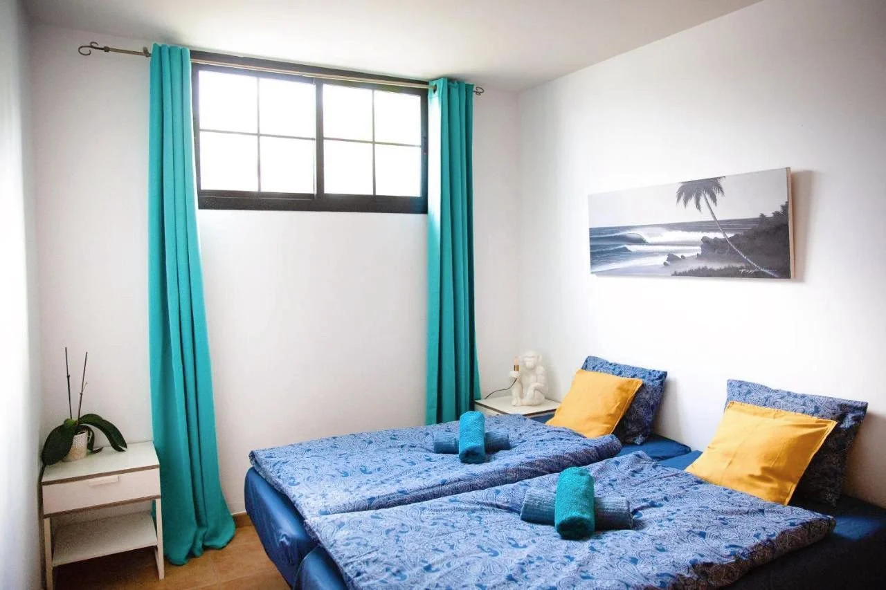 A bedroom with two beds, blue and yellow pillows, blue bedspreads, turquoise curtains, a framed black and white seascape photograph on the wall, and a window with black framing.