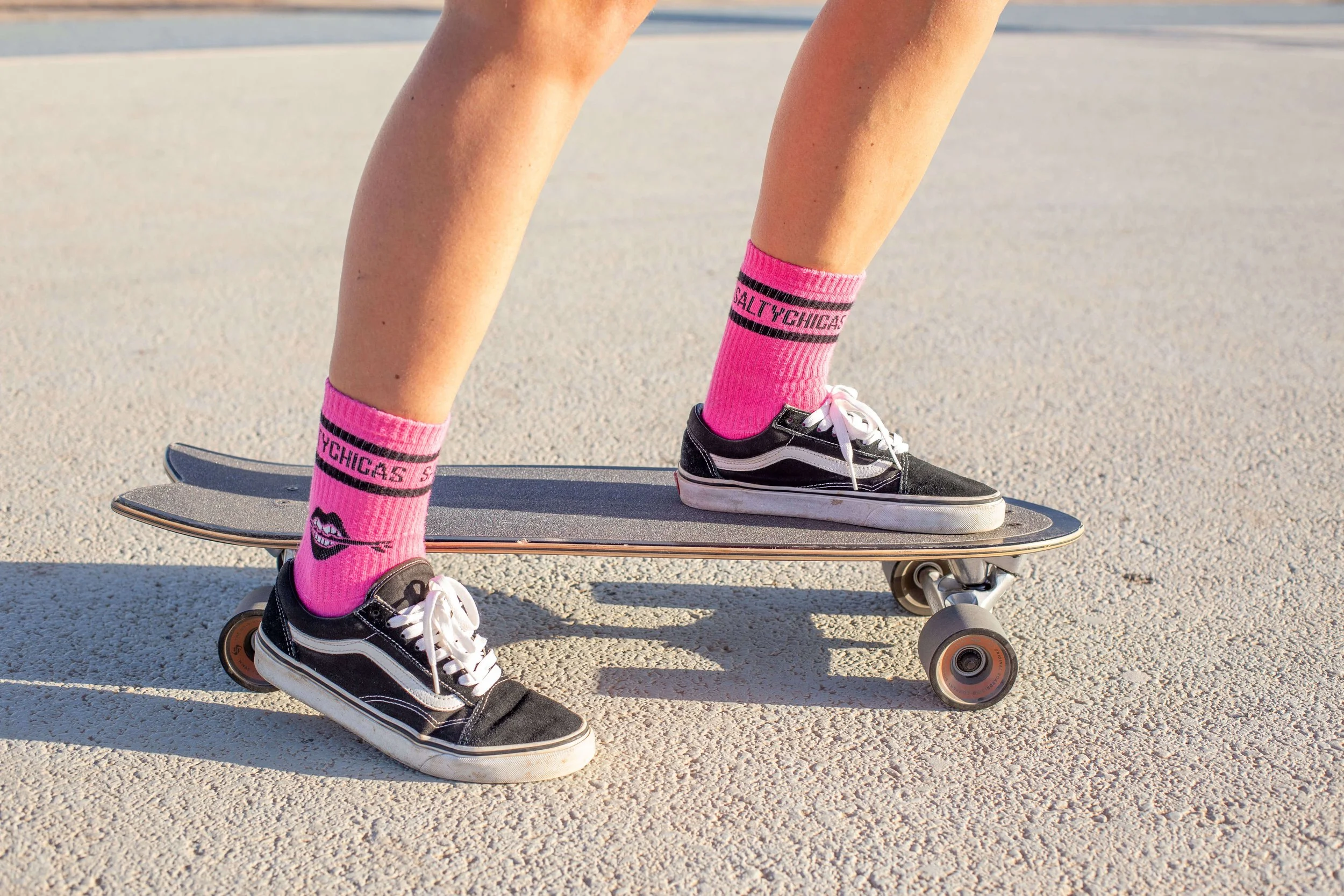 Person standing on skateboard wearing black Vans sneakers and bright pink socks with the text and face design, on a sandy surface.