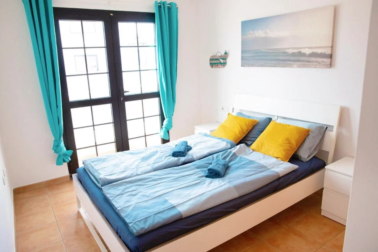 A bright bedroom with a black-framed glass door, turquoise curtains, white walls, a beach-themed painting, a white bed with yellow pillows, and neatly folded towels on the bed.