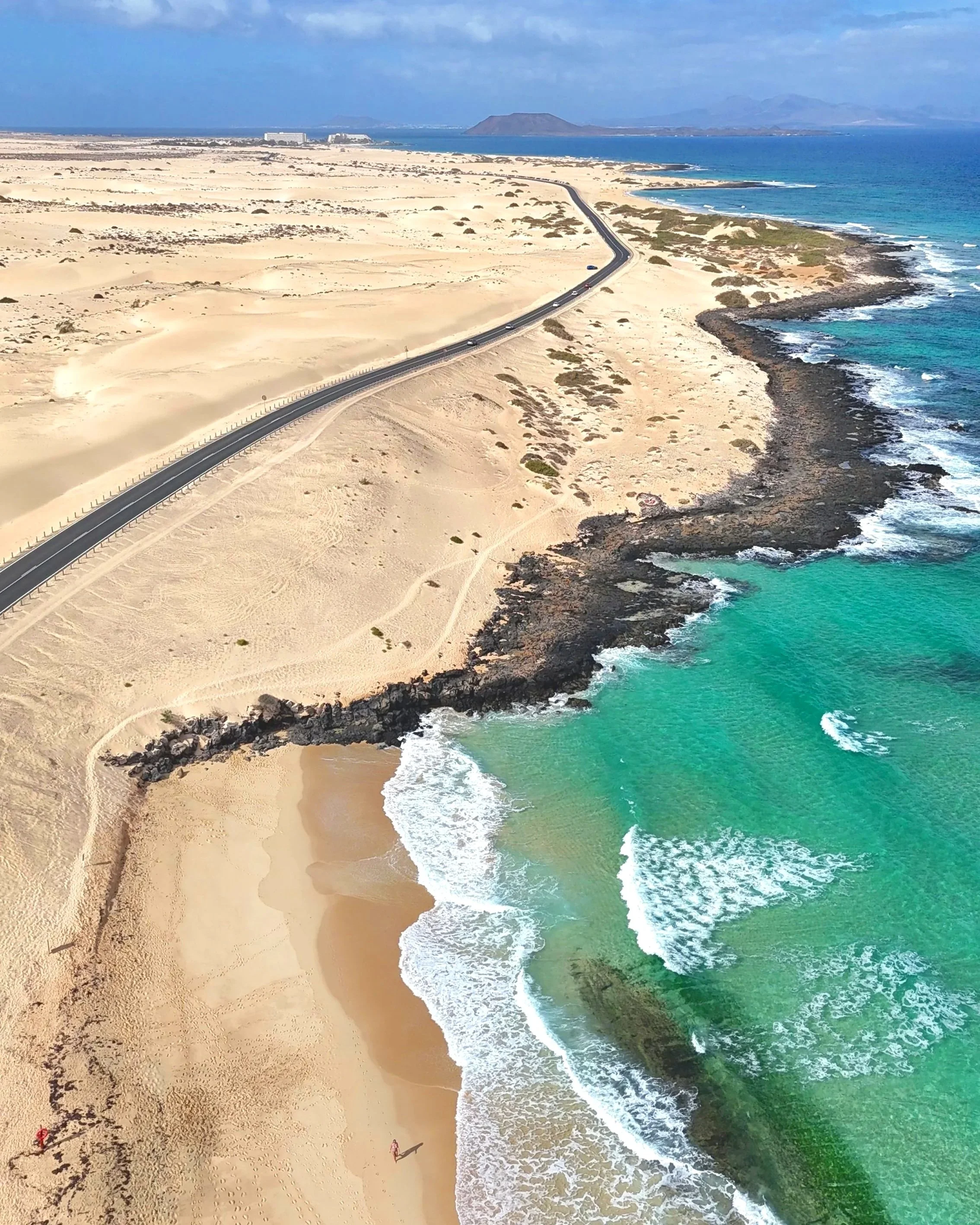 Aerial view of a beach with turquoise water, black rocks, sandy shore, and a road running parallel to the coastline in a desert landscape.