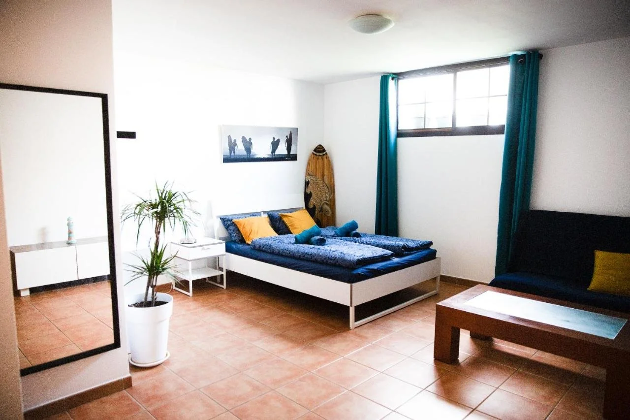 A bedroom with a large bed with blue bedding, yellow pillows, and rolled towels, a surfboard, a black sofa with a yellow pillow, a potted plant, a rectangular mirror, and a window with teal curtains.