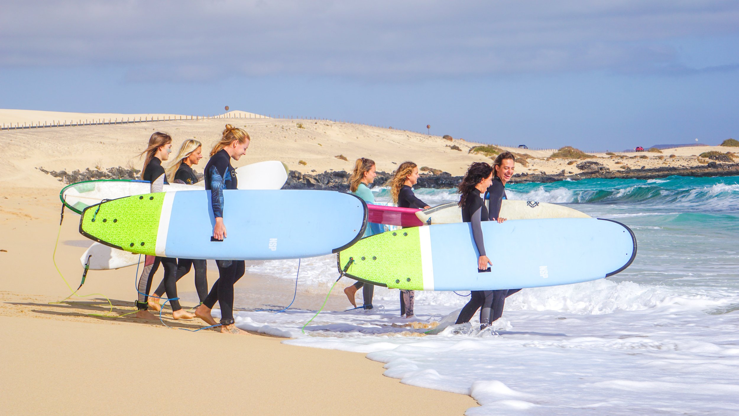 A group of women carrying surfboards walking into the ocean on a sandy beach, with waves and a cloudy sky in the background.