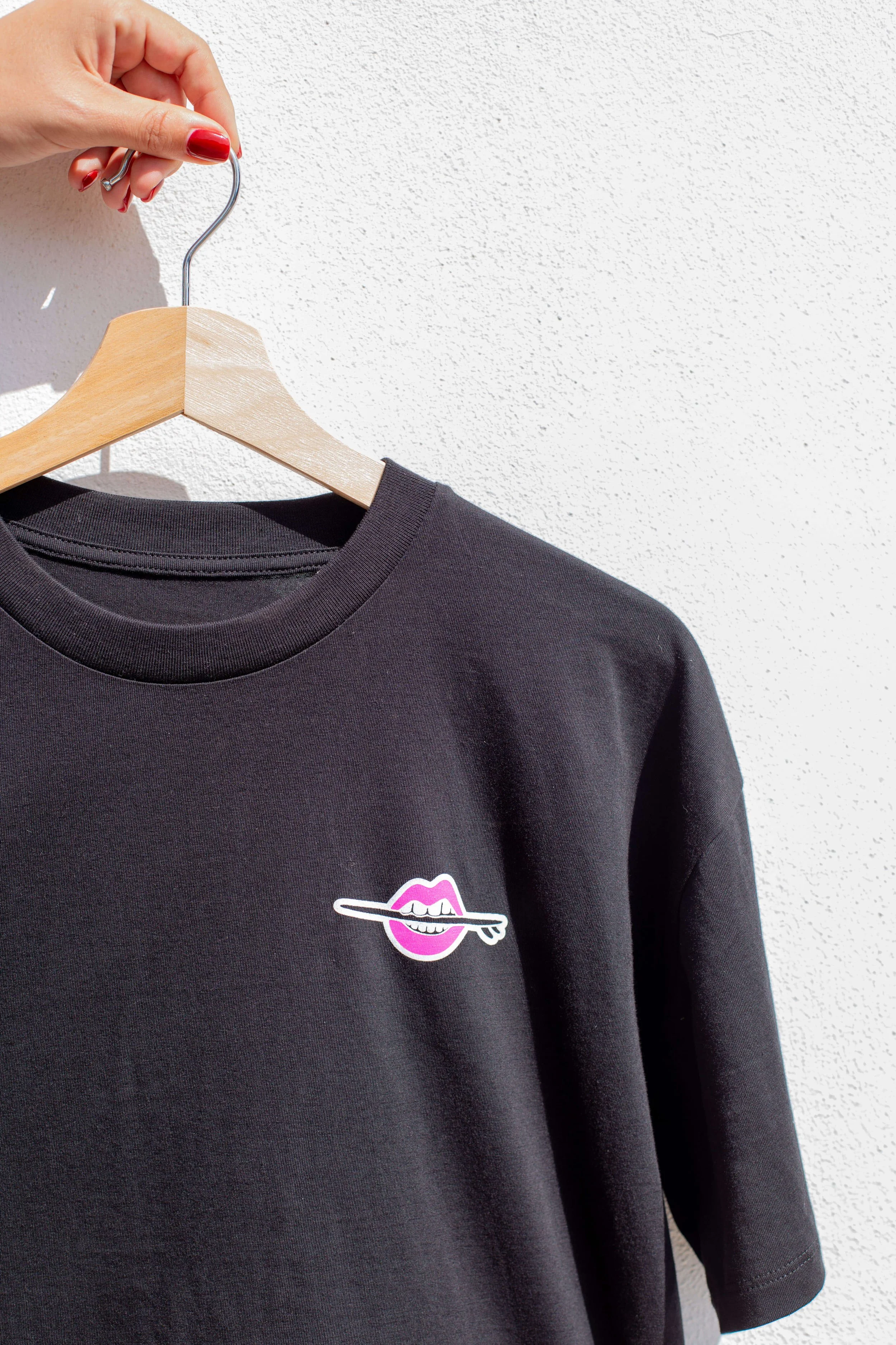 A black sweatshirt with a graphic of pink lips and a white line, hanging on a wooden hanger against a white textured wall.