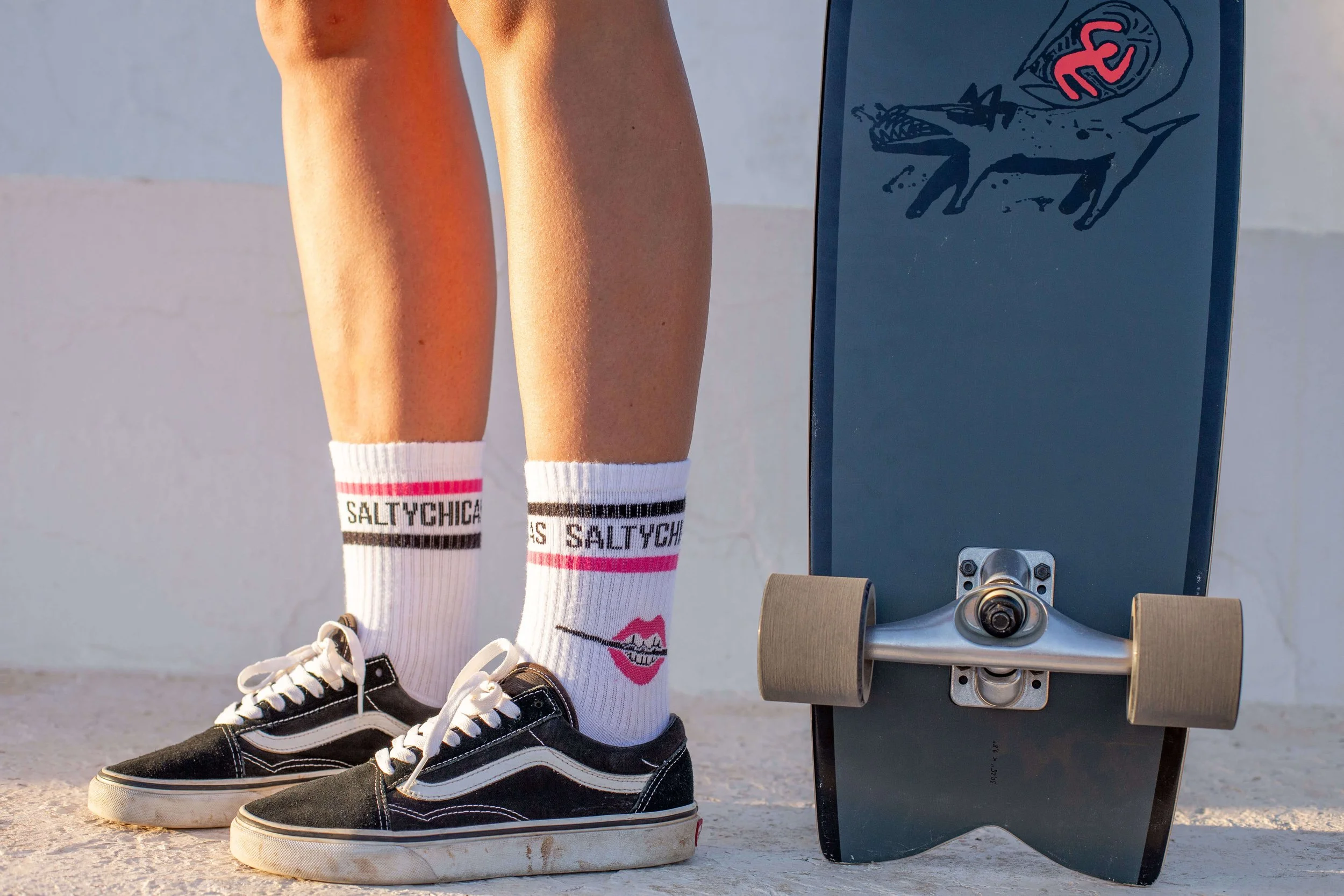 Close-up of a person wearing black Vans sneakers and white Salt Y Chicago socks with pink and black stripes, standing next to a skateboard with a blue deck and beige wheels, against a white wall.