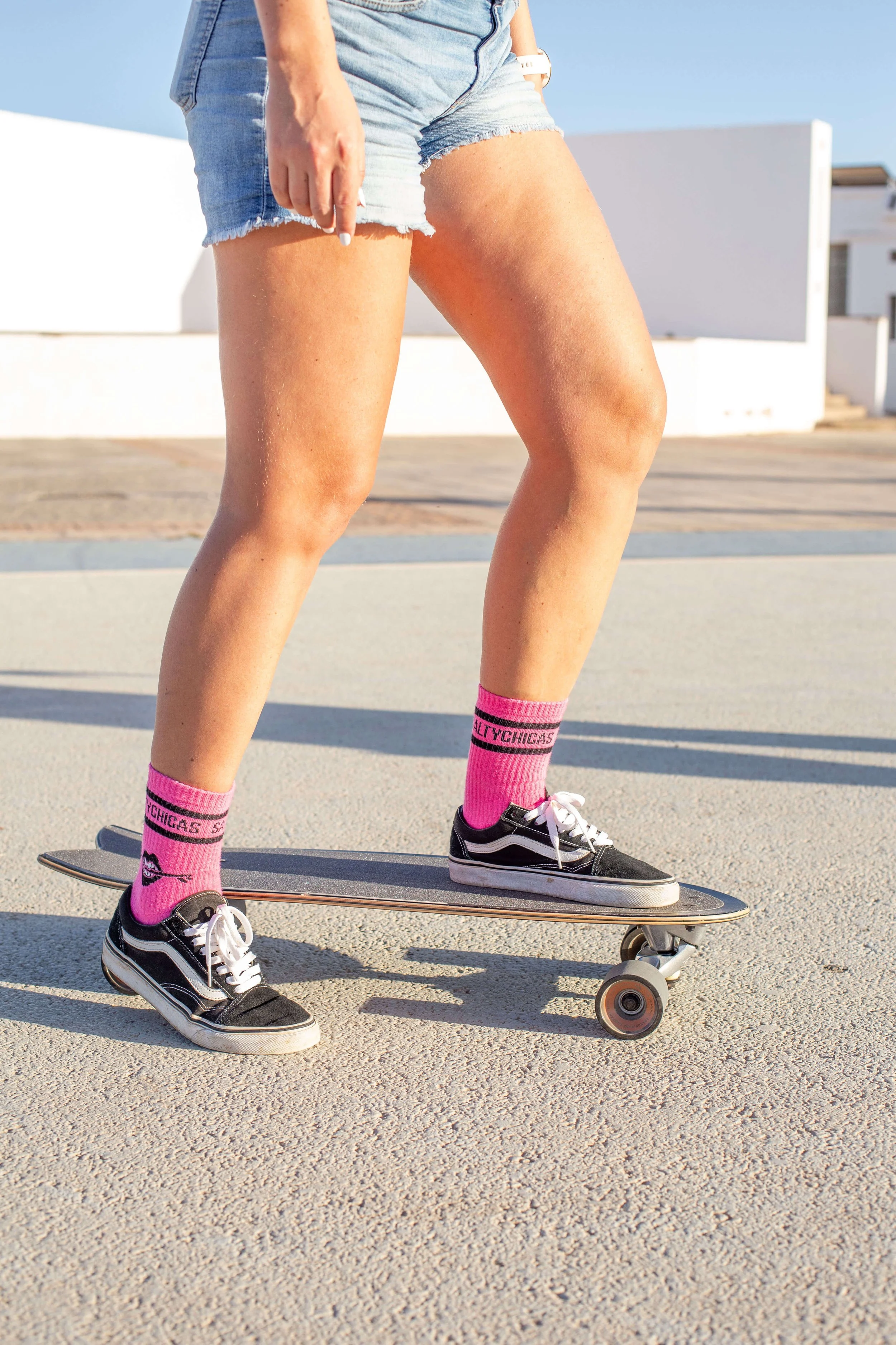 Close-up of a person wearing denim shorts, pink socks, and black sneakers riding a skateboard on a concrete surface outdoors in sunny weather.