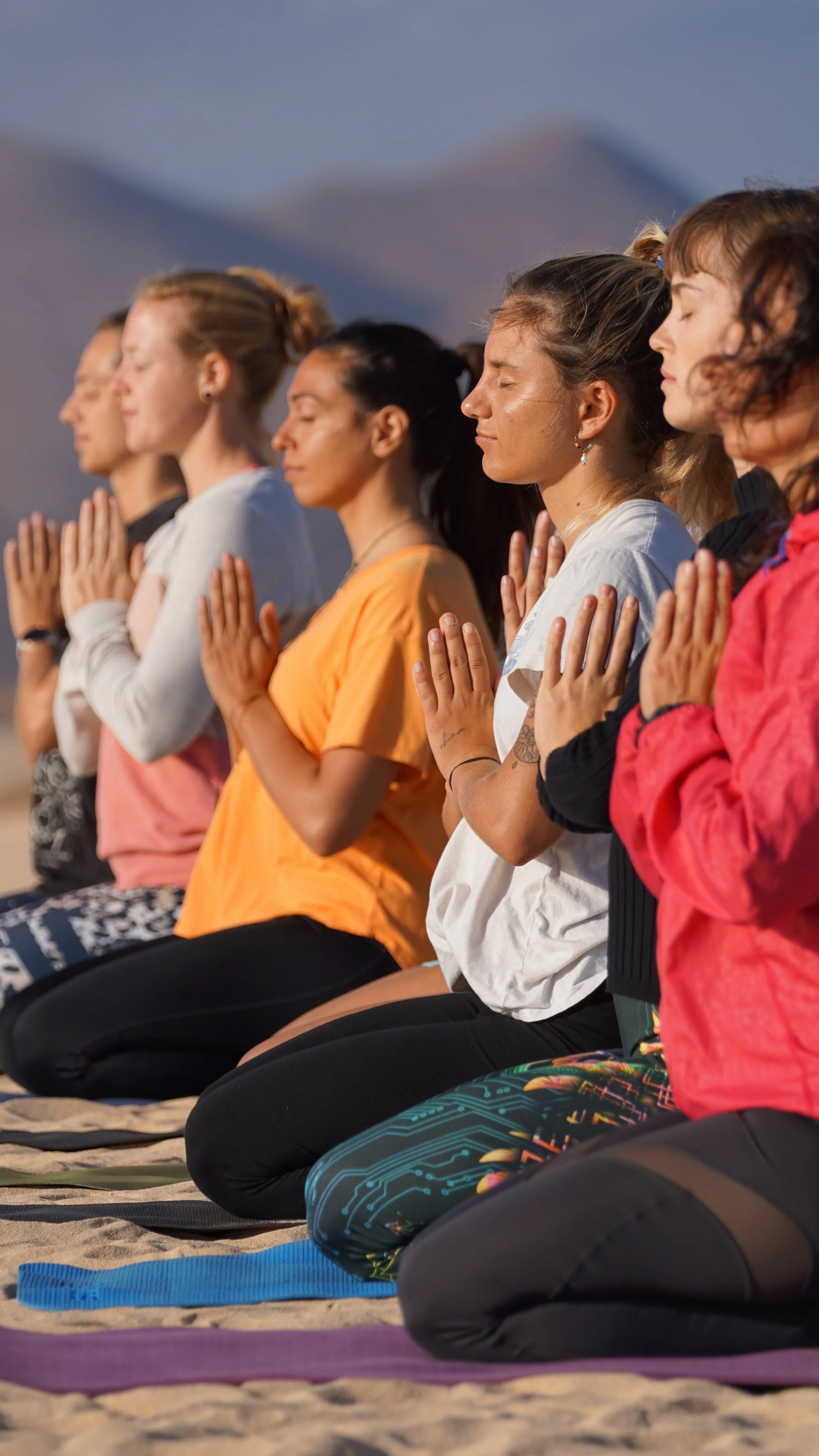 A group of women practicing yoga outdoors on a sandy surface, sitting in a kneeling meditation pose with eyes closed.