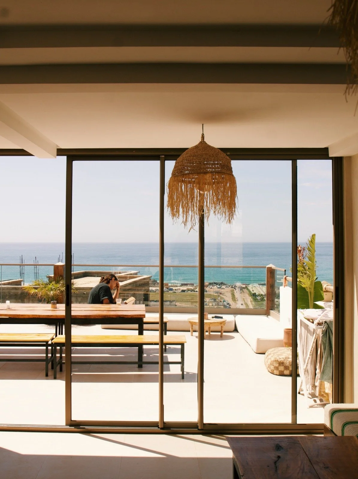 Living room with large glass sliding doors leading to a balcony overlooking the ocean, with a woman reading at a wooden table, and a hanging wicker lamp in the ceiling.
